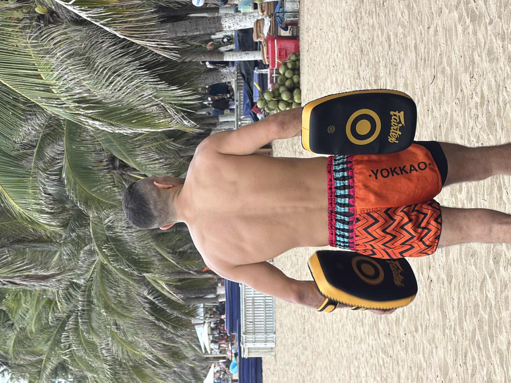 A shirtless man with a close-cropped haircut stands on a sandy beach in Nha Trang, Vietnam, facing away from the camera. He is wearing orange Muay Thai shorts with a black and teal zigzag pattern and a black under-layer. The man is holding two black and yellow Muay Thai kick pads, one in each hand. In the background, palm trees line the beach, and further beyond, several people are visible near white tents and blue seating. A pile of coconuts and red cooler boxes are also present on the sand.
