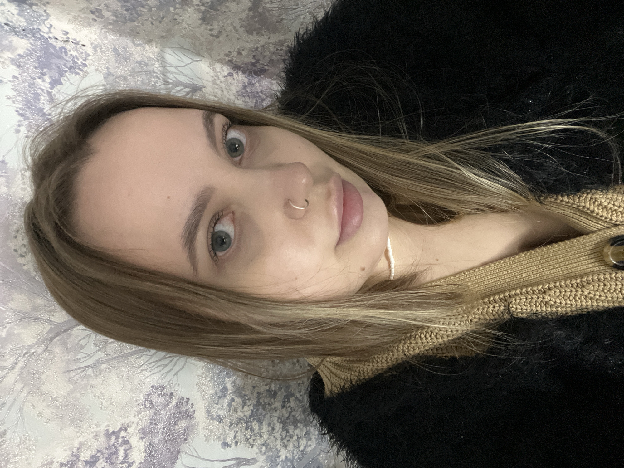 A young woman is pictured in a close-up selfie. Her hair is light brown with blonde highlights, and she has fair skin, green eyes, and a nose ring. She is wearing a black, fuzzy jacket over a tan sweater. A white beaded necklace is visible around her neck. The background is a patterned surface, possibly a bedspread or wall. The location is not discernible from the image. The image does not provide enough information to determine that it was taken in Novopokrovskaya, Russia.