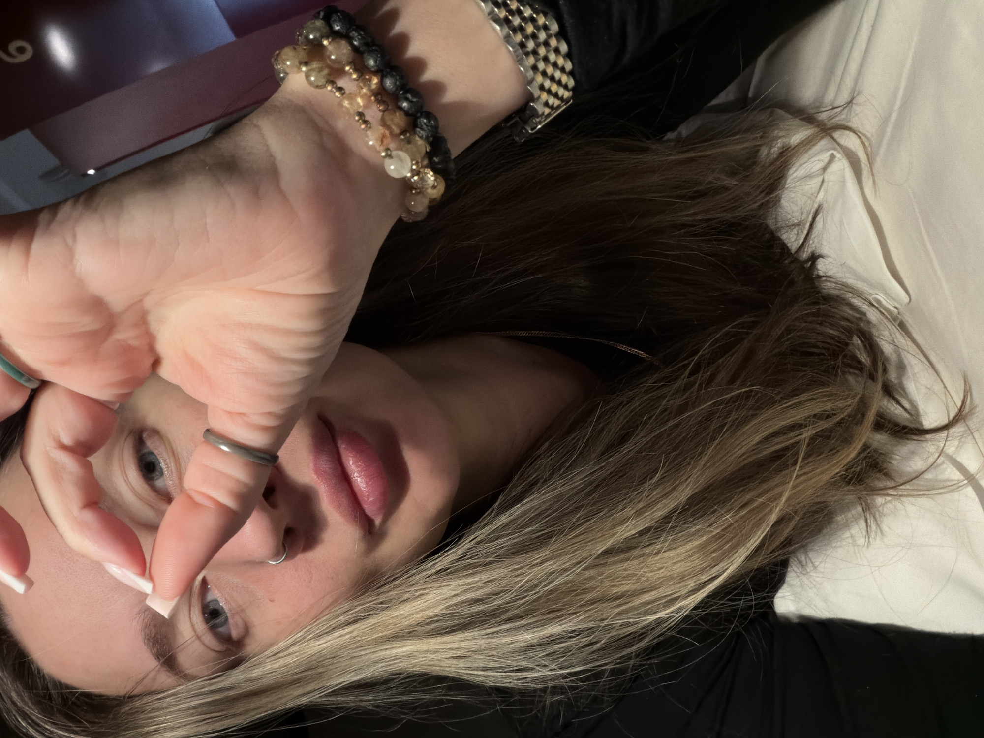 An angled, close-up portrait features a person, likely female, lying down indoors. The individual's head is tilted, with their blue eyes looking directly at the viewer through an opening formed by the fingers of a raised hand. The hand partially covers the face, and a silver ring adorns the ring finger, while the fingernails display a French manicure. The person's lips are a natural pink, and a silver hoop nose ring is visible.
Long, light brown hair with blonde highlights cascades over a white surface, possibly a pillow or bedding. The person is wearing a black garment. On the visible wrist, multiple beaded bracelets are stacked, composed of various materials, including dark, porous beads and lighter, translucent, golden-toned beads. A silver-toned watch with a checkered metal band is also worn.
The background is dimly lit, with a soft reddish-pink glow and a partial view of the number "6" in the upper left corner, suggesting an indoor setting like a room or lounge. The overall mood is relaxed. While the image itself contains no specific geographical markers, it is provided within the context of Moscow, Russia.