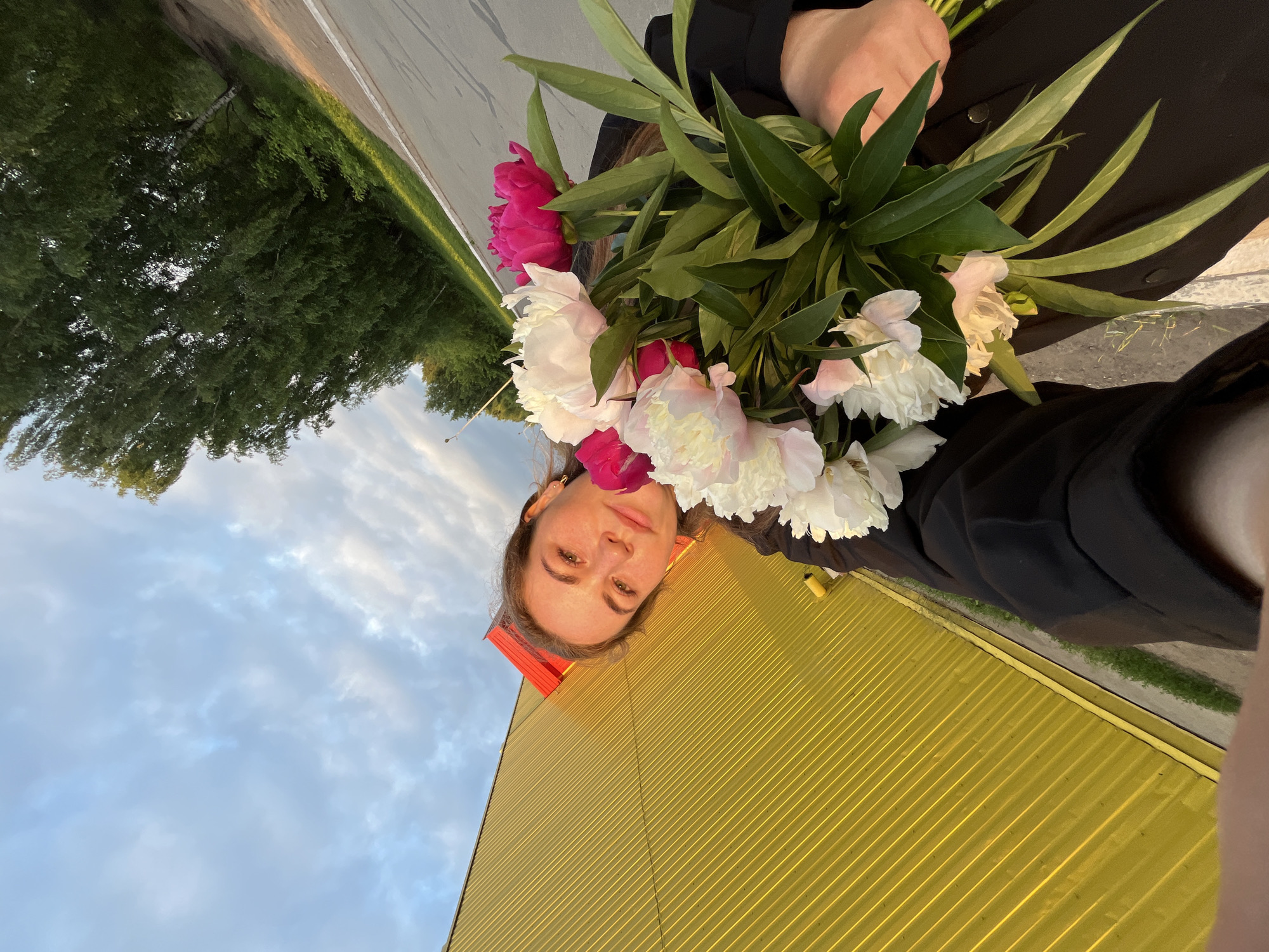A young woman is taking a selfie outdoors, holding a bouquet of peonies. She is wearing a black jacket. The background shows a bright yellow building with corrugated siding, a paved area, trees, and a cloudy sky. The image appears to have been taken in the city of Chaikovsky, Russia, possibly near an industrial or commercial area, given the building's appearance. The lighting suggests it is either early morning or late afternoon.