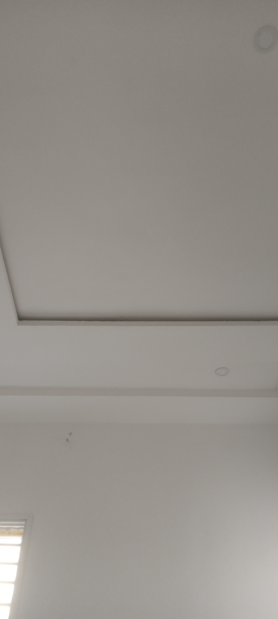 The image shows a portion of an interior space, likely a room, with a focus on the ceiling and the upper part of a wall. The ceiling features a rectangular recessed section, and two recessed lighting fixtures are visible. The wall is painted white. A portion of a window is visible in the lower left corner, suggesting natural light is entering the room.