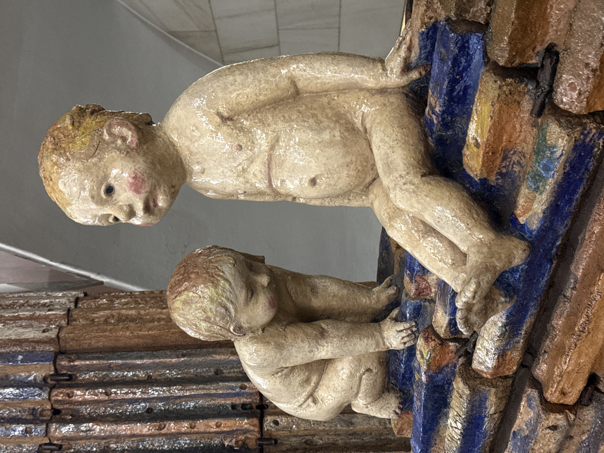 This media file, captured in Moscow, Russia, displays a close-up of a decorative architectural element featuring two cherubic, doll-like figures clinging to a textured, multi-colored structure. The figures, appearing to be infants or toddlers, are rendered in what looks like glazed ceramic or terracotta, showcasing a pale, somewhat glossy skin tone with visible textural details.
The larger, upper figure is positioned vertically, facing downwards and slightly to the left. Its face features delicate sculpting with dark eyes and rosy cheeks, and faint indications of hair. Its nude torso and limbs exhibit a slightly lumpy, handcrafted appearance, and its feet are shown gripping a prominent blue vertical section of the background.
Below it, a smaller figure actively clutches a similar blue vertical element with both arms wrapped around it. This figure's head is tilted downwards, creating a sense of concentration or effort in its clinging posture. Both figures convey a placid, unexpressive demeanor, typical of such decorative art.
The background structure behind the figures is composed of rough, block-like elements, alternately colored in vivid blue and earthy orange-brown tones. These elements create a rugged, uneven surface. In the lower left of the image, behind the smaller figure, several rows of dark, textured, horizontal components are visible, possibly representing roof tiles or another section of an architectural facade. The scene is brightly and evenly lit, emphasizing the handcrafted textures and vibrant colors of the artwork. No text is visible in the image.