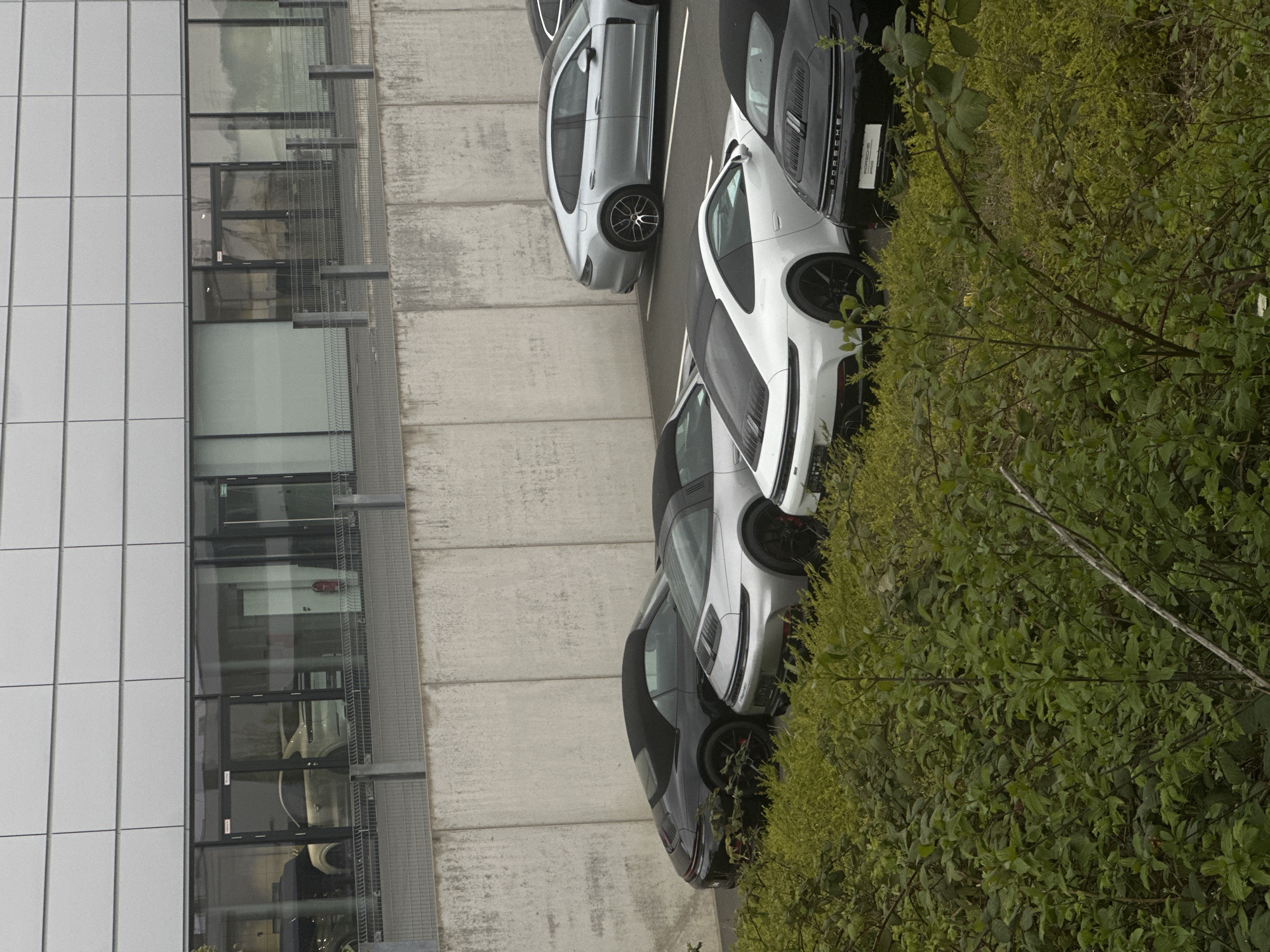 The image is an angled aerial shot of a scene in Holzwickede, Germany. A modern building with a grid-like facade of white panels and dark-framed windows dominates the left side of the image. To the right of the building, a concrete-paved parking area contains several luxury cars, including what appear to be Porsches, parked in a row. The cars are various colors, including silver, white, and black. Dense green foliage and bushes occupy the right side of the image, partially obscuring the cars and adding a natural element to the urban scene. The overall composition suggests an upscale or corporate environment with high-end vehicles.