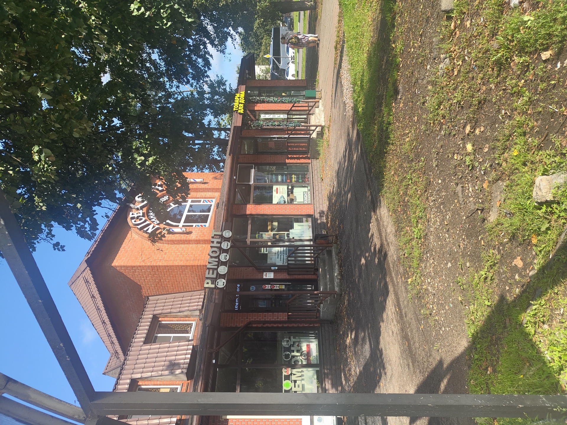 The image shows a brick building in Ramenskoye, Russia, with a storefront that appears to be a repair shop and a tobacco shop. The building has a brown tiled roof with a dormer window. Above the repair shop entrance, there is a sign with the word "РЕМОНТ" (REPAIR) in large letters. Next to the repair shop is a sign for a tobacco shop that reads "ТОВАРЫ ДЛЯ ДОМА" (GOODS FOR HOME) in yellow letters. The storefront windows display various items, including what appear to be phone cases and accessories. A barber pole symbol is visible on the building's upper section. The area in front of the building is paved with concrete and a sidewalk. Trees with green leaves are present on the right side of the image, and a small patch of grass and dirt is in the foreground. In the background, a bus and several cars are visible on a street. Two women are walking on the sidewalk in the middle ground. The sky is clear and blue.