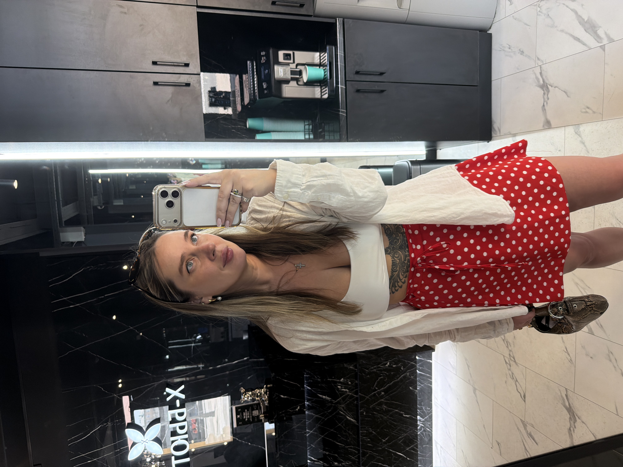 A woman is taking a selfie in what appears to be a salon or retail space. The woman has light brown hair, blue eyes, and is wearing a white top, a red skirt with white polka dots, and brown shoes. She has a tattoo on her upper side. She is holding a smartphone with a clear case. In the background is a black countertop with a glossy finish and a cabinet area above it. The salon logo "TOPOL-X" is visible on the countertop. The location is likely Moscow, Russia. The floor has a white marble-like pattern. There are reflective surfaces throughout the space, including what appears to be a mirror behind the woman.