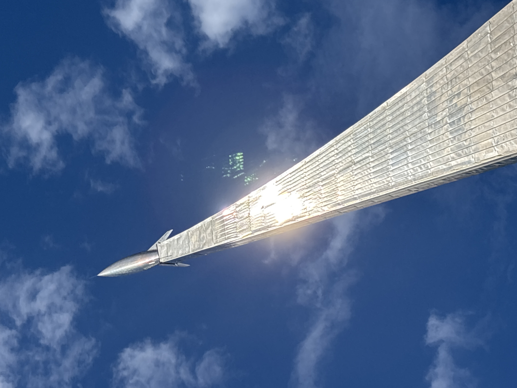 The image shows a low-angle view of the Monument to the Conquerors of Space in Moscow, Russia, set against a blue sky with scattered clouds. The monument, made of a shiny metal resembling titanium, appears to be reflecting sunlight. The monument's rocket is visible at the lower left. The sun's reflection creates a bright glare on the monument, obscuring some of the details.