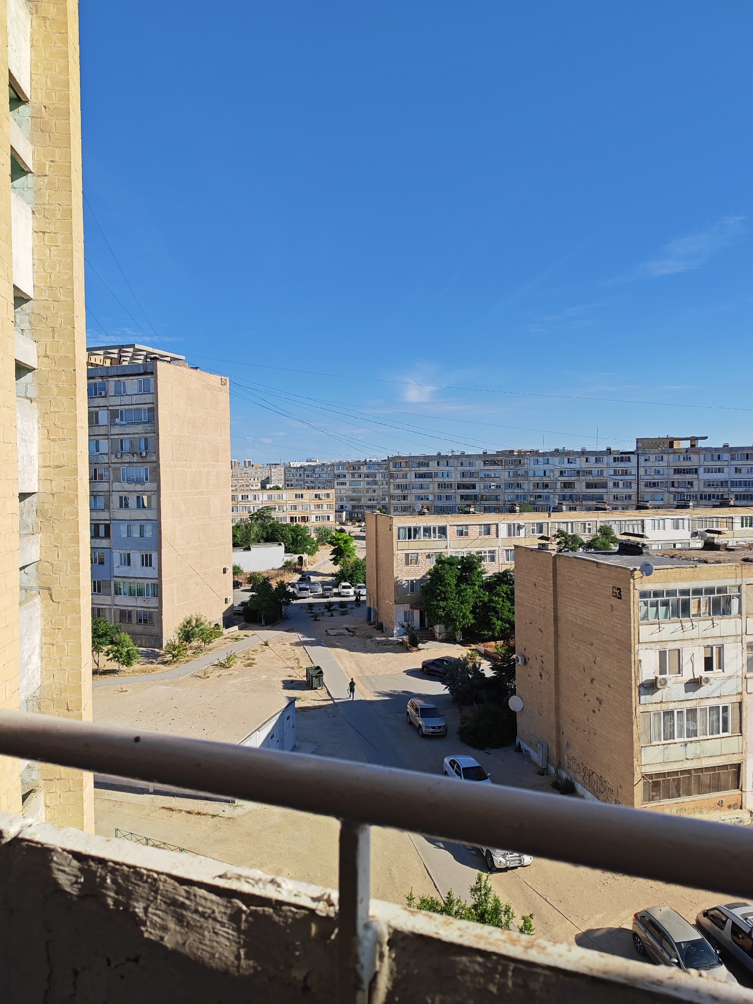 This is an outdoor, eye-level, medium shot of an urban landscape under a clear blue sky. In the foreground, a rusty metal railing runs horizontally across the lower part of the frame, partially obscuring the view. To the left, a tall building facade made of yellow brick with white trim dominates the left edge of the image.
The mid-ground reveals a street lined with several multi-story apartment buildings. These buildings appear to be constructed from a combination of beige and white materials, with numerous windows visible. Some of these buildings exhibit signs of wear and tear, with peeling paint and visible air conditioning units. Trees and shrubs are scattered throughout the area, with a particularly dense patch of green visible behind one of the beige buildings. Several cars are parked along the street or driving. A dark-colored sedan is parked on the left side of the street, and a light-colored SUV and a white sedan are driving on the road. A single person is walking along the sidewalk in the middle distance, near a large green trash bin.
In the background, a long, flat, multi-story apartment building stretches across the horizon, punctuated by many windows. The overall impression is that of a residential area, possibly a Soviet-era development, characterized by functional architecture and a somewhat weathered appearance. The setting is consistent with the appearance of residential areas in Aktau, Kazakhstan, a coastal city known for its distinctive architectural style developed during the Soviet period. The clear blue sky suggests it is a sunny day.