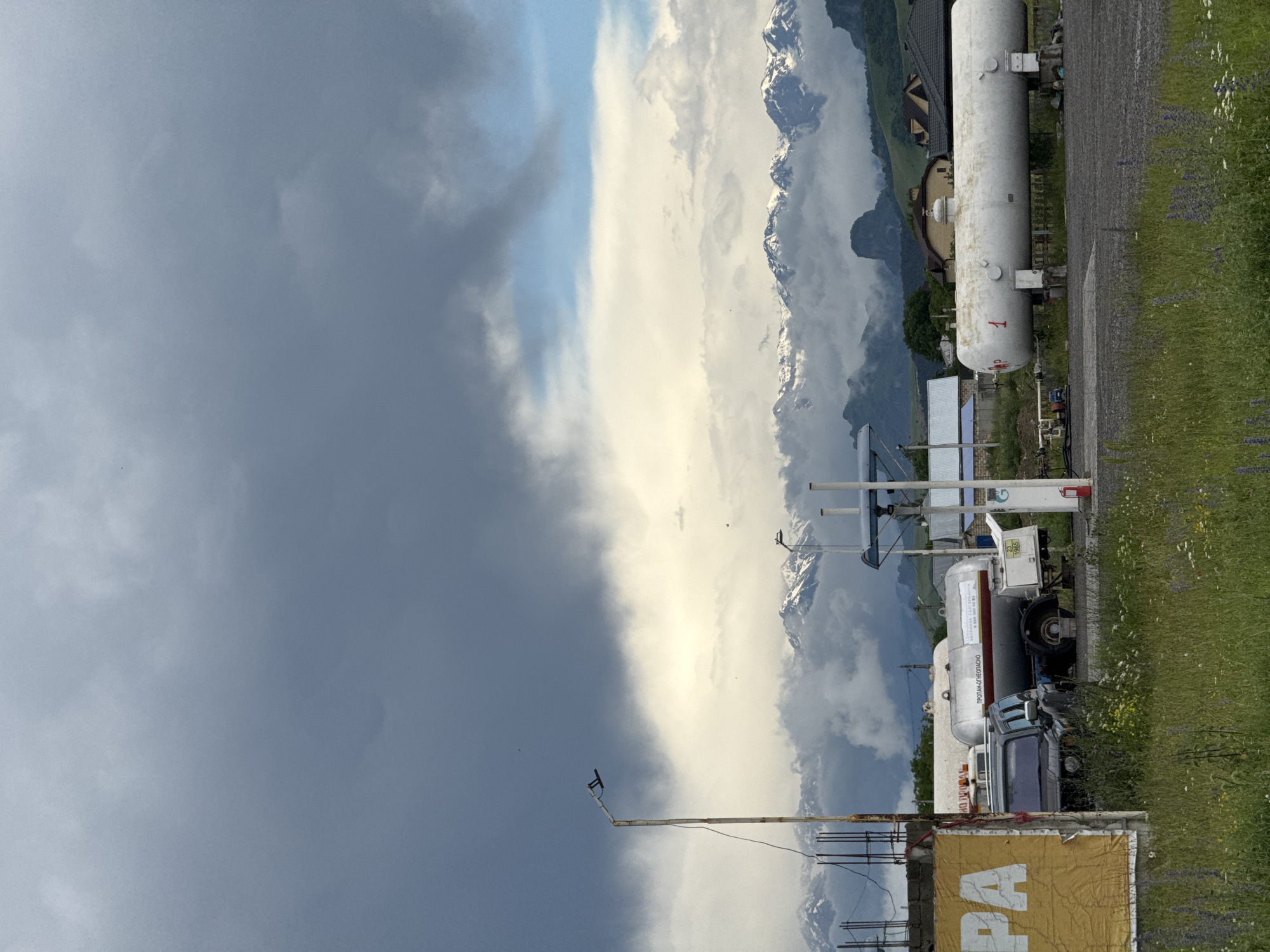 The image depicts an outdoor scene in Kumysh, Russia, featuring a landscape with prominent mountains in the background, partially obscured by clouds. In the foreground, there's a gas station with several large white propane tanks, one of which is labeled with the number "1". A truck, likely a tanker, is parked near the tanks. A dark-colored SUV is also visible. A street lamp stands to the left of the frame, and a yellow sign with partial lettering is present on the left side. The ground is covered with gravel and green grass, with some wildflowers. The sky is filled with dramatic, dark storm clouds contrasted with patches of brighter sky. The overall impression is of a rural or semi-industrial setting against a dramatic natural backdrop.