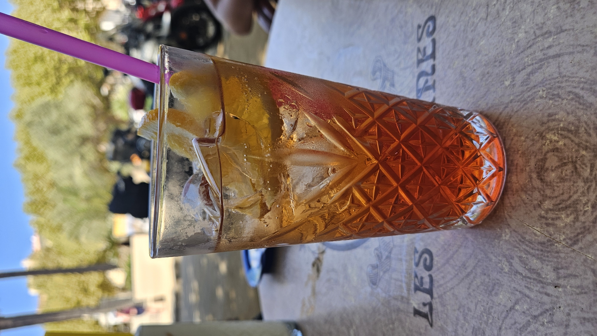 A tall glass filled with an orange beverage, ice, and orange slices sits on a patterned table outdoors. A pink straw extends from the glass. In the background, blurred trees and buildings suggest a sunny day. The table appears to be at a cafe or restaurant in L'Île-Rousse, France, as evidenced by the partial lettering "LES" on the table surface, likely part of a restaurant name or signage. The overall scene is casual and suggests leisure.