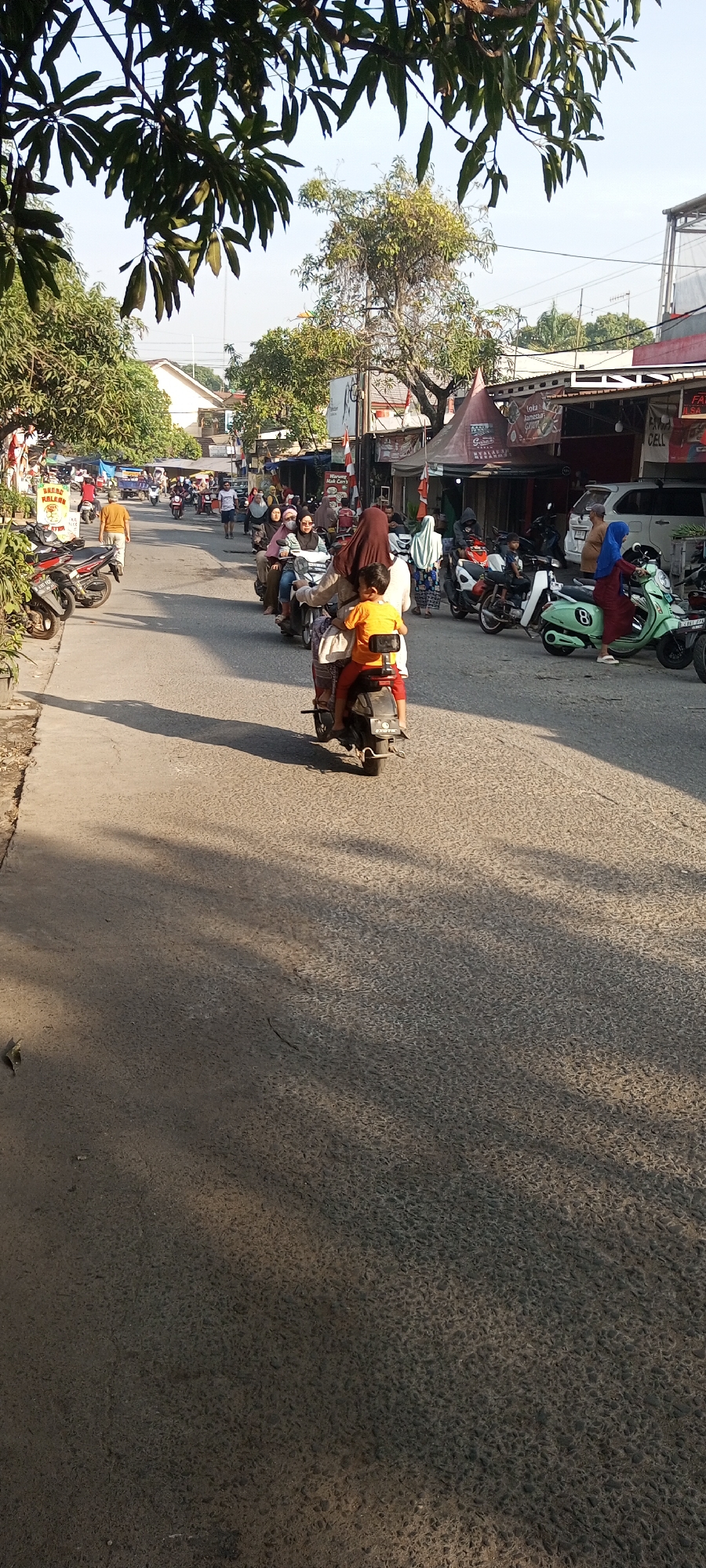 The image shows a street scene in Setu, Indonesia, under a bright, hazy sky with sunlight casting shadows on the asphalt road. A procession of motorcycles dominates the foreground, with riders and passengers, mostly wearing headscarves, navigating the street. One prominent motorcycle carries a child in an orange shirt in the front, with an adult riding behind. To the left, several parked motorcycles are visible. The background features a row of buildings, some with awnings and signage, and lush green trees. The overall impression is of a busy, sunlit day on a local street in Indonesia, likely a market or commercial area given the presence of shops.
