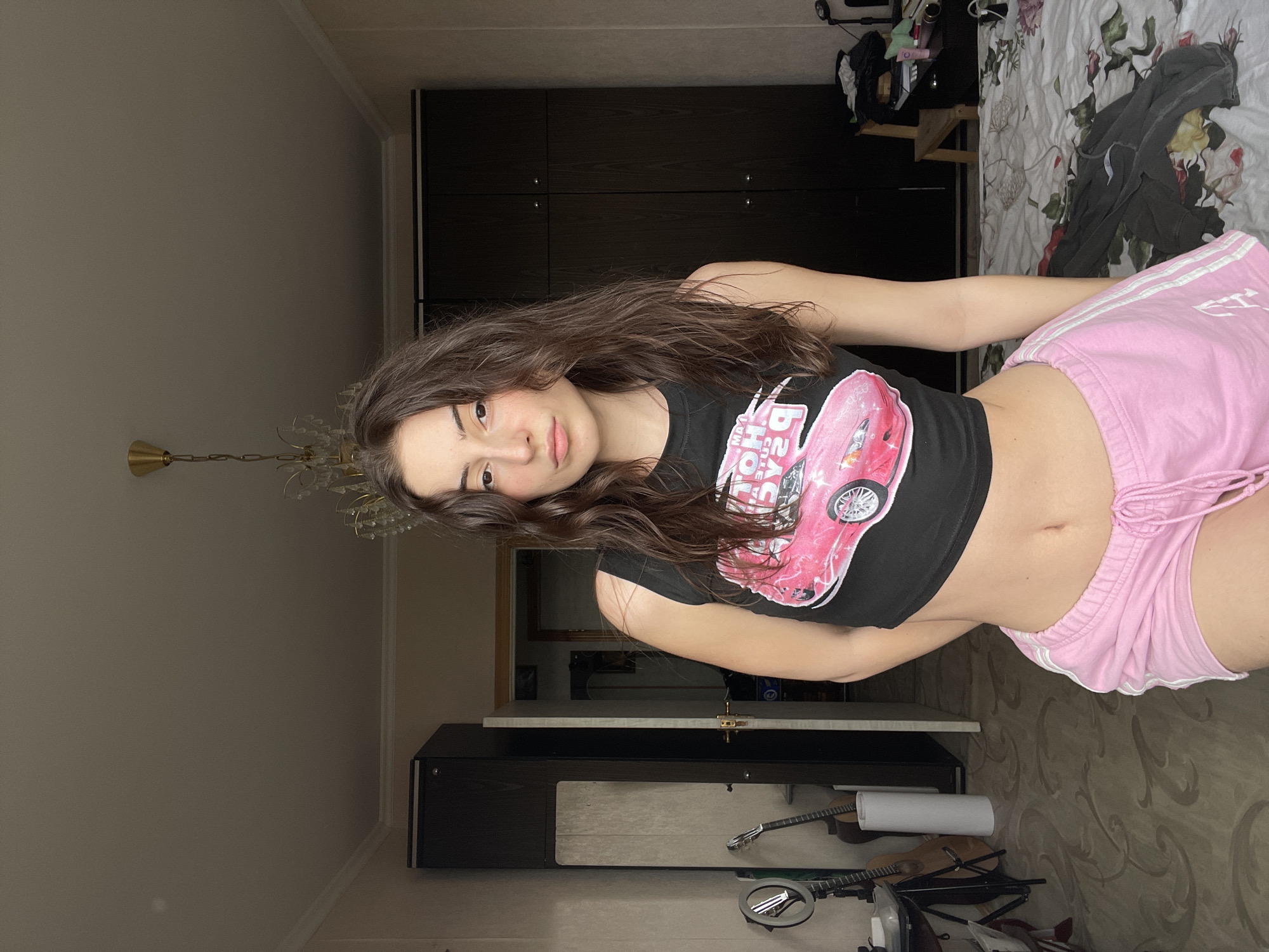 The image depicts a young woman with long, dark hair, wearing a black crop top with a pink car design and pink shorts. She is indoors, likely in a bedroom or living space, as suggested by the presence of a bed with patterned bedding, a dark wardrobe, and a chandelier. A desk and musical instruments are also visible. While the image is taken indoors and could be located anywhere, there is no visual information to definitively place the scene in Moscow, Russia.