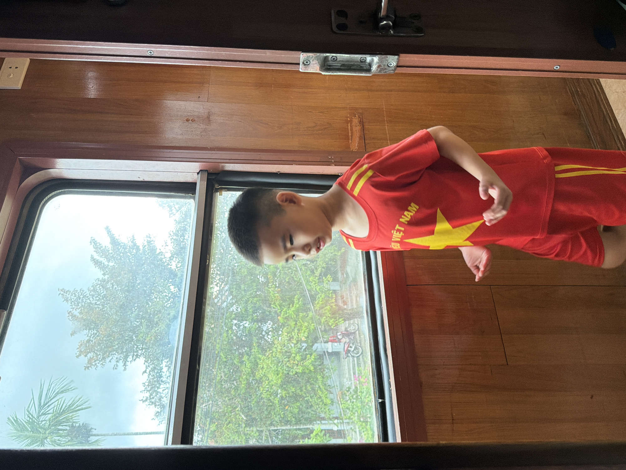 A young boy wearing a red t-shirt and shorts with yellow trim, featuring a yellow star and the text "VIỆT NAM", stands inside a train carriage, looking out of the window. The interior of the carriage is paneled with wood. Outside the window, trees and a motorbike are visible under a cloudy sky, suggesting a location in Đà Nẵng, Vietnam. The scene captures a moment of observation from within the moving train.