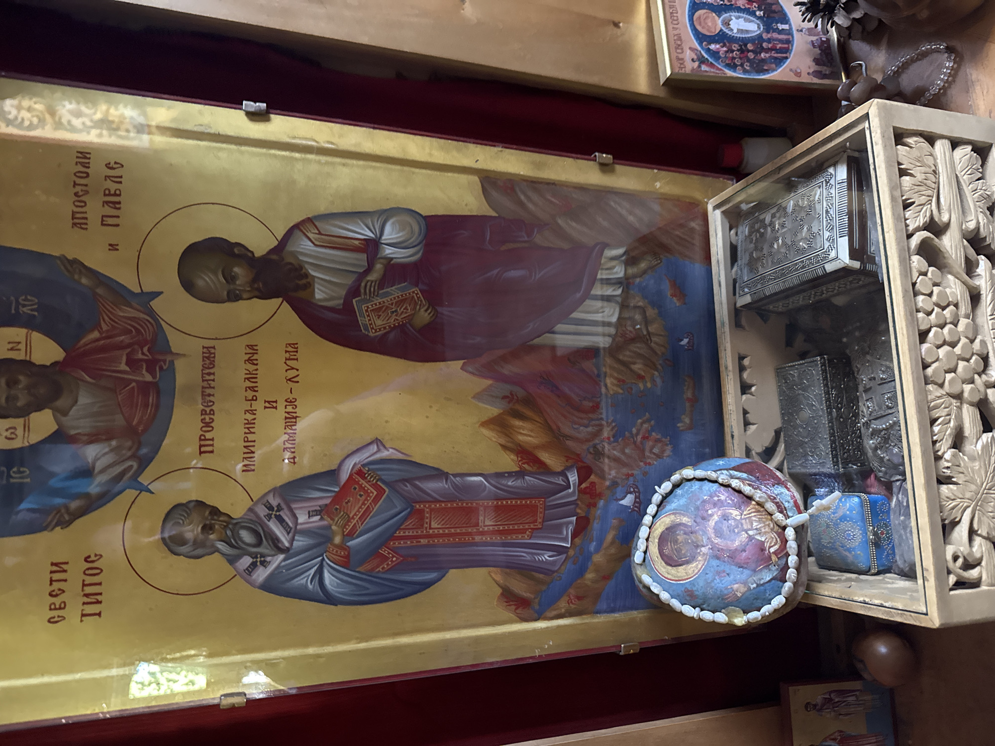 The image displays an icon of Saint Titus and Saint Paul in a room, possibly a church or a private collection, with religious artifacts. The icon, gold-leafed and framed, depicts Saint Titus on the left, wearing a blue and purple robe and holding a red book, and Saint Paul on the right, in a white and maroon robe, also holding a book. Inscriptions in Cyrillic are visible above and beside the figures.
In the foreground, a carved wooden box contains several items. A round, painted stone with an image of the Virgin Mary and Child, adorned with a string of light-colored beads, rests on top of the box. Inside the box, there are at least two ornate metal boxes and a smaller blue decorative box. To the left of the carved box, a small framed icon is visible, and further left, a round, brown object, possibly a fruit or a decorative item, sits on a surface. To the right of the carved box, a small pinecone and prayer beads are visible, along with another framed icon on the far right, showing a circular depiction of figures surrounding a central figure in white. The background is red and gold.
This scene is located in the Monastery of Tvrdoš in Trebinje, Bosnia and Herzegovina.