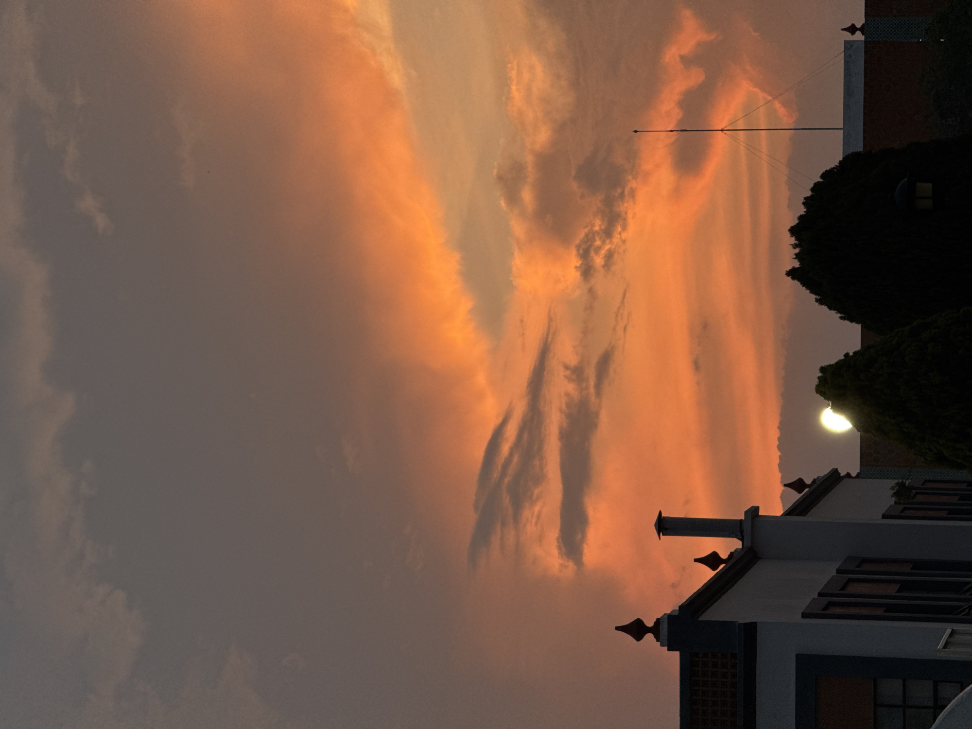 The image depicts a sunset in San Andrés Cholula, Mexico. The sky is filled with orange and gray clouds. In the lower right of the frame, the corner of a light-colored building with dark window trim is visible. A chimney and decorative finials are also present. To the right of the building are dark green trees. A tall antenna is seen against the sky, reaching over a dark building. The moon is visible on the right of the frame. The general atmosphere appears to be dusky and quiet.