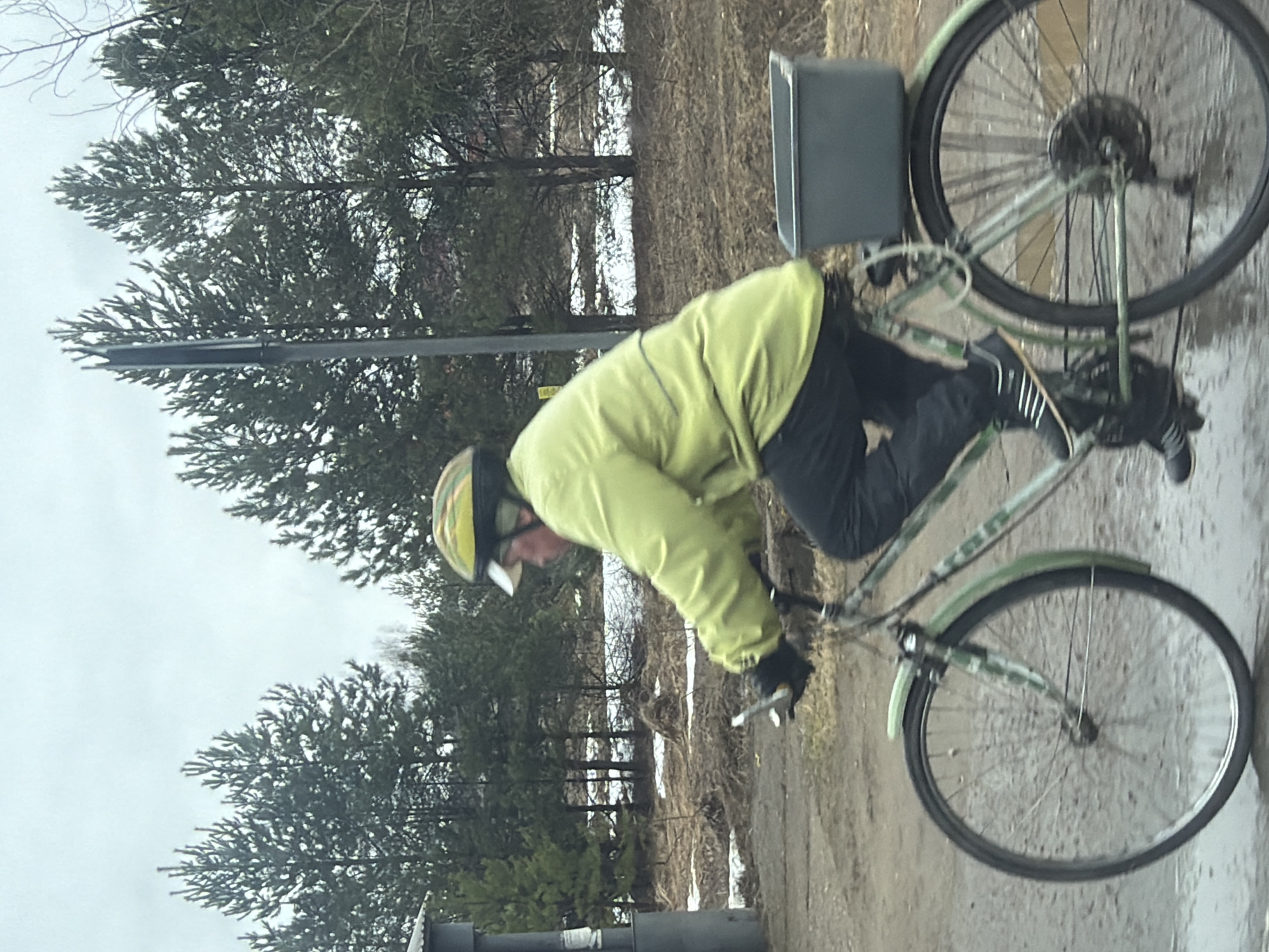 A person wearing a yellow jacket, black pants, and a helmet is riding a green bicycle on a muddy path. The bicycle has a basket on the back. The location appears to be near a forest with snow patches and a utility pole. Based on the prompt, the location is Прикамский, Russia.