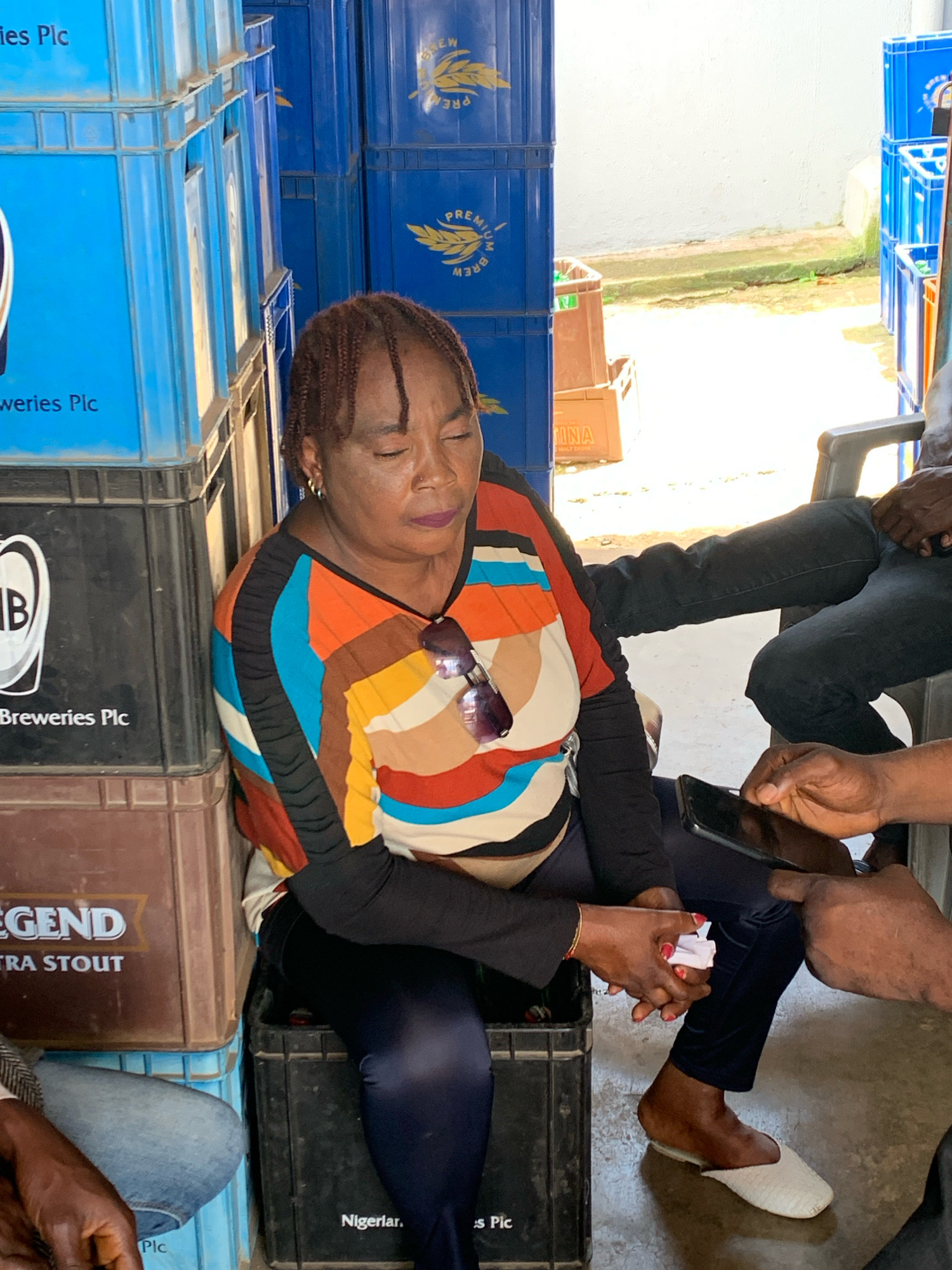 A middle-aged Black woman with braided hair is seated on a stack of black plastic crates, with her eyes closed and head tilted slightly. She is wearing a long-sleeved black top with a colorful striped pattern across the chest and abdomen, and dark blue leggings. A pair of sunglasses is resting on her chest. Her left hand is holding a small white object, possibly paper, and her right hand is resting on her lap. Beside her, a Black person in black jeans and a dark shirt is seated on a chair. Another Black person's hands are visible on the right, holding a smartphone and pointing at it. The background consists of stacks of blue plastic crates, some with "PREMIUM BREW" labels and wheat imagery, and brown crates with "LEGEND EXTRA STOUT" labels. The scene appears to be an outdoor or semi-outdoor area, possibly a storage or distribution point, in Jos, Nigeria, suggested by the "Nigerlan. s Pic" visible on one of the crates. The ground is a concrete surface.