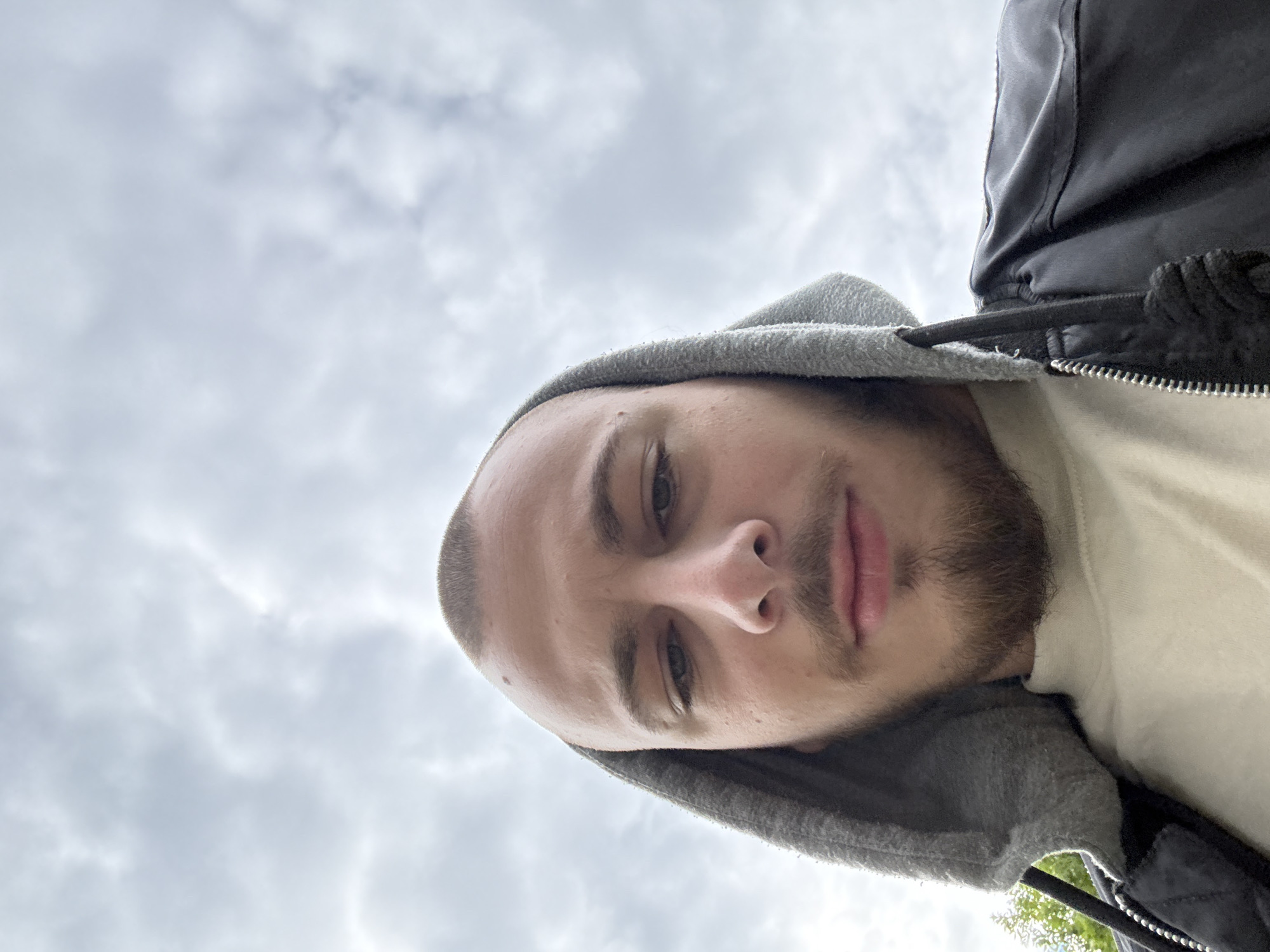 The image is a rotated close-up selfie of a young man, capturing his face and upper torso against an overcast sky. The man has short, possibly buzzed hair and a short beard or stubble covering his chin and jawline. He looks directly into the camera with a neutral expression, revealing light-colored eyes and a defined nose and lips.
He is wearing a grey hooded sweatshirt with the hood pulled up, layered beneath a dark, likely black, jacket with a visible silver zipper on the right side of the frame. The material of the grey hood appears somewhat textured.
The background is predominantly a light grey, cloudy sky, indicating an outdoor setting during daytime in Stara Zagora, Bulgaria. The clouds are diffuse and spread across the sky. A small glimpse of green foliage, likely from trees, is visible in the bottom right corner of the image, peeking from behind the man's jacket. The photograph was likely taken with the camera held at a low angle, pointing upwards towards the subject. No text is visible in the image.