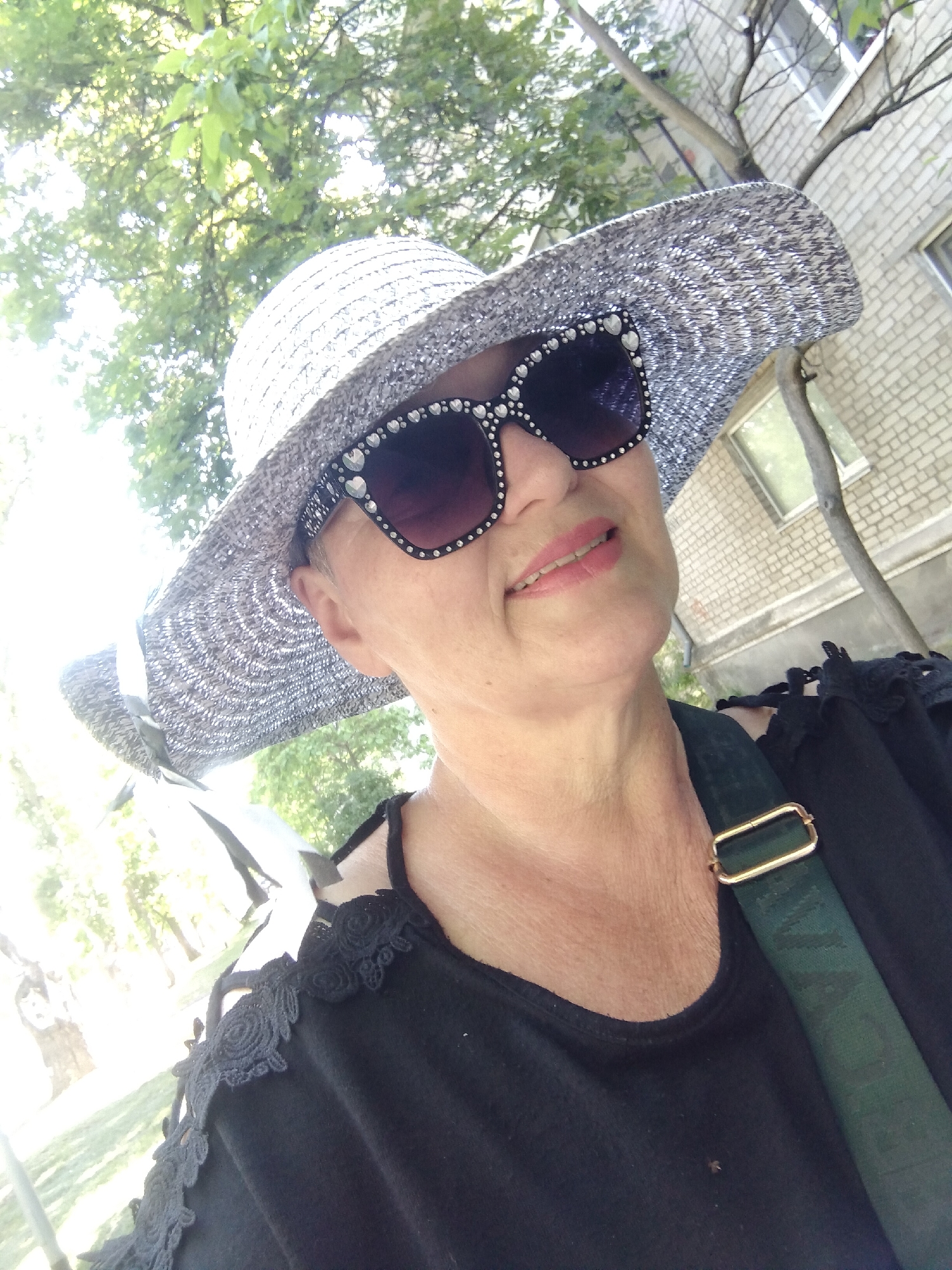 A woman, wearing a wide-brimmed, sparkly hat and ornate sunglasses, poses for a selfie outdoors. She has light skin and is wearing a black top with lace trim and a green strap with a gold buckle across her chest. The background shows trees with green leaves and a brick building. The location is the city of Horishni Plavni, Ukraine.