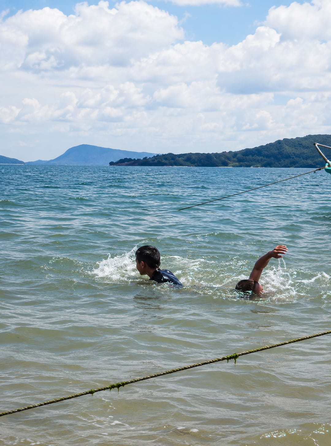Two young boys are swimming in the water near the shore of Lumban Bul Bul, Indonesia. The boy on the left is wearing a dark blue shirt and is facing to the right, with his head turned towards the right. He is creating a splash of water as he swims. The boy on the right is mostly submerged, with only his head and right arm visible above the water. His right arm is extended upwards, and he appears to be reaching for something. The water is choppy and appears murky. In the background, there is a body of water stretching out to a distant shoreline lined with green trees and hills. The sky is blue with scattered white clouds. A rope is visible in the foreground, stretching across the water. There is also a hint of a green object, possibly part of a boat or buoy, to the right of the frame.