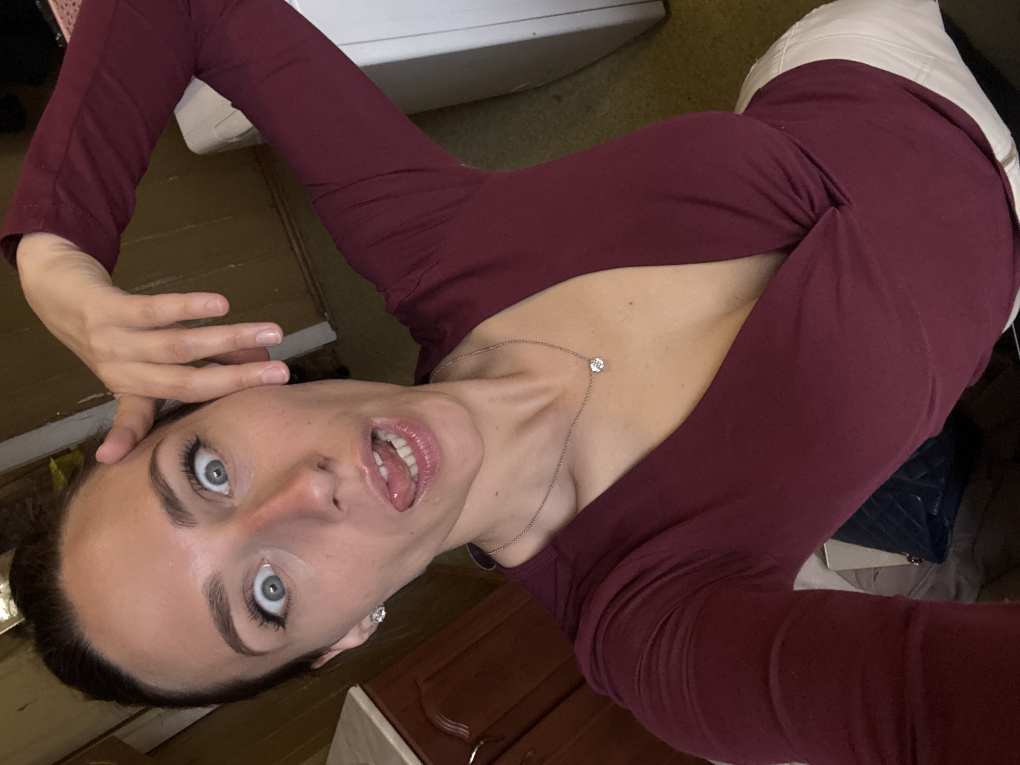 A young Caucasian woman with dark hair and blue eyes is posing for a selfie. She is wearing a maroon long-sleeved shirt and white pants. She has a silver necklace with a diamond pendant and a small earring in her left ear. She is holding her left hand near her face and sticking out her tongue. The background includes a wooden floor, white furniture, and dark wooden cabinets. The location appears to be indoors. There is no evidence to suggest this photograph was taken in the city of Saint Petersburg, Russia.