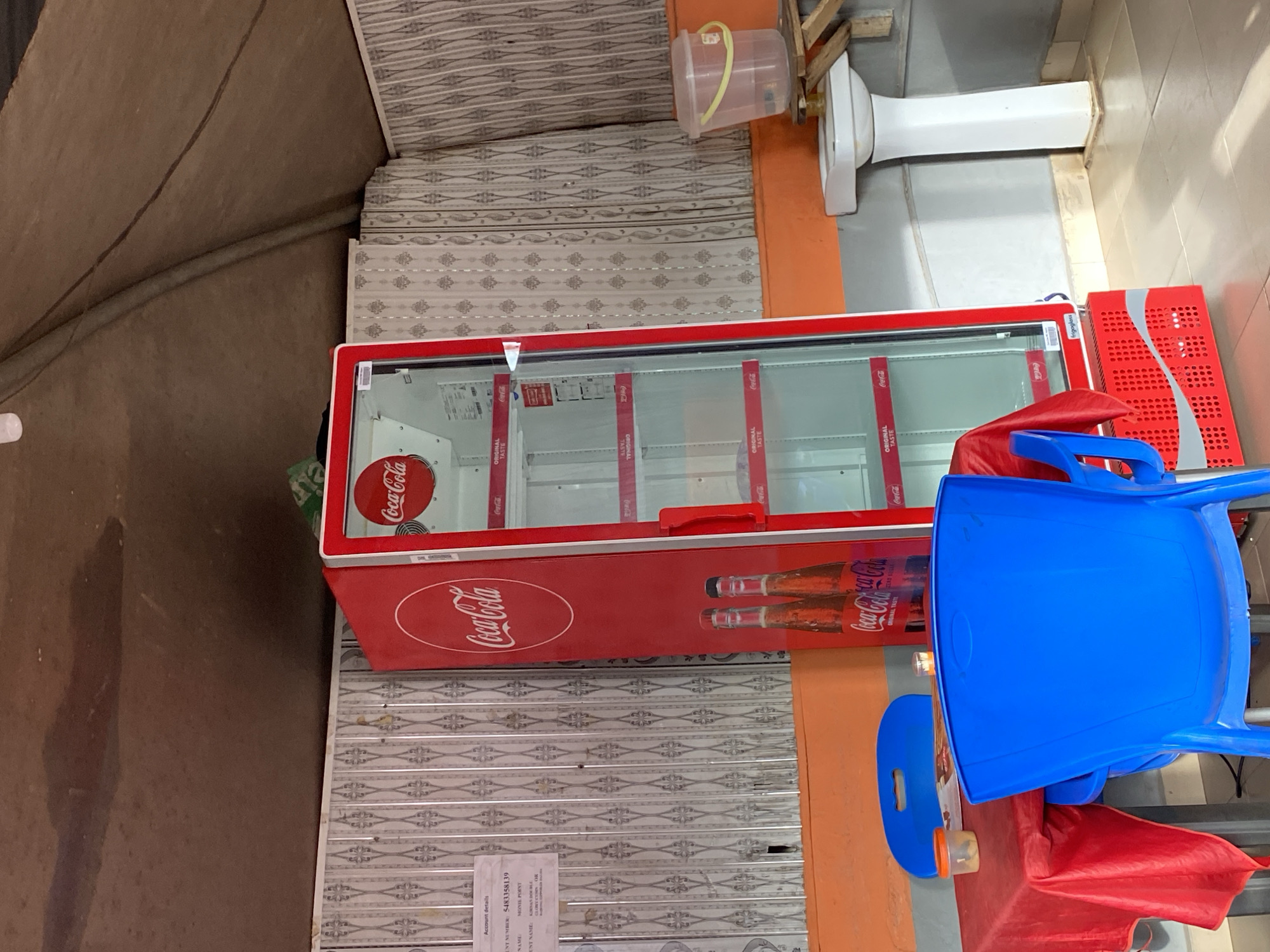 This media file, oriented sideways, captures a scene likely from a small commercial establishment in Jos, Nigeria. The central subject is a large, red Coca-Cola branded display refrigerator, which appears to be empty, suggesting it might be closed, awaiting restock, or out of service. The cooler features the iconic "Coca-Cola" logo prominently on its front and also on internal dividers, which additionally display the text "ORIGINAL TASTE." The brand name "frigoglass" is visible on the side of the cooler.
To the right of the cooler, there are two bright blue plastic chairs and a red plastic Coca-Cola crate. Further right, a white, toilet-like fixture, possibly a hand-washing station or water dispenser, is mounted on an orange-colored wall. Above it, a clear plastic container with a yellow handle is suspended.
The background consists of patterned grey and white roller shutters or panels, possibly the front of the shop or a ceiling feature. A brown wall or canvas canopy covers the upper left portion of the image.
On the lower left, affixed to the patterned panels, a printed paper displays "Account details," including "ACCOUNT NUMBER: 5483358139," and partially visible text such as "ACCOUNT NAME: MODOX POINT" and "CURRENT CUSTODIA." No people are visible, and the time of day is not discernible.