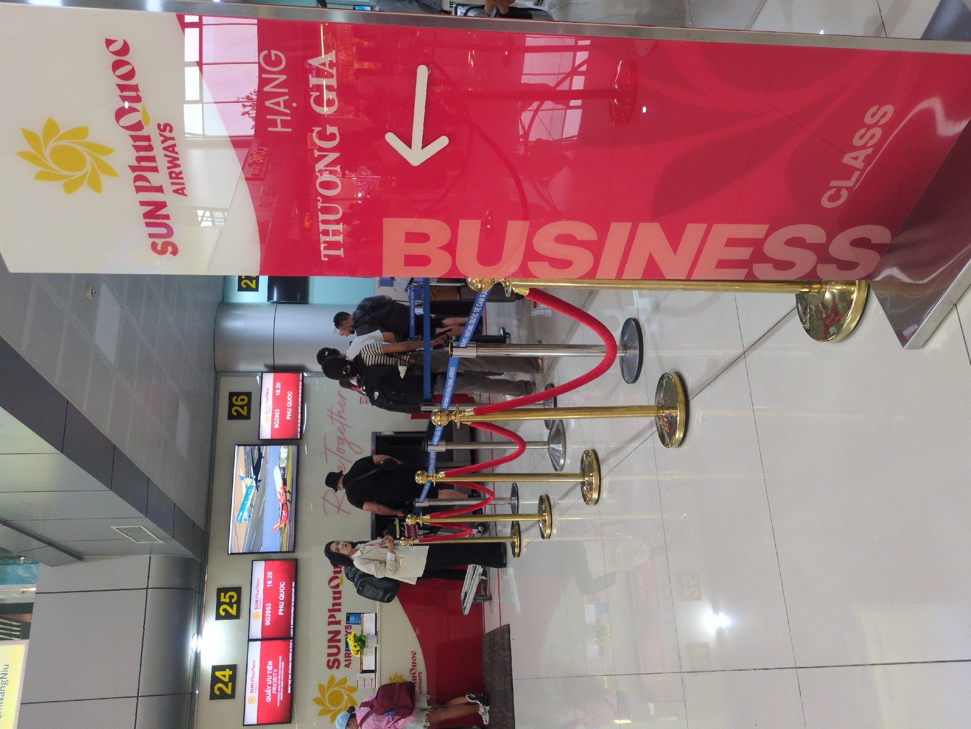 The image depicts a business class check-in area at an airport, likely in Cam Ranh, Vietnam, given the presence of Vietnamese text on signs. The scene features a queue of people waiting to check in, demarcated by red velvet ropes and gold stanchions. A prominent red sign with the words "BUSINESS CLASS" and "THƯƠNG GIA" (meaning business class in Vietnamese) hangs above the queue. The sign also displays the logo of Sun Phu Quoc Airways. In the background are flight information screens displaying flight details for Phu Quoc destinations, including flight number 9G2953. Check-in counters are visible behind the queue, with staff assisting passengers. The floor is light-colored tile.
