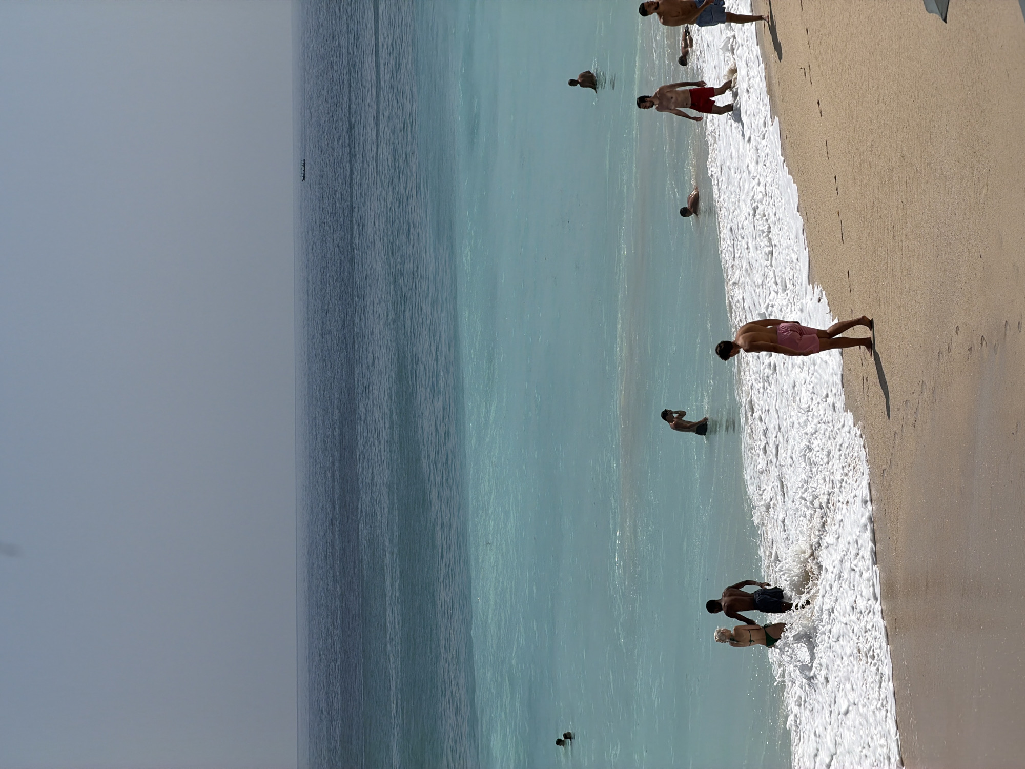 The image shows a beach scene in Kuta Selatan, Indonesia. The ocean is a vibrant turquoise with a line of white surf breaking on the sandy shore. Several people are visible, some in the water and some on the beach. In the foreground, a man in pink shorts walks along the sand. Further back, a couple stands in the shallow surf, surrounded by white foam. Other individuals are scattered throughout the water, swimming or wading. The sky is a hazy, pale blue, and a small boat is visible in the distance on the dark blue expanse of the ocean. The overall impression is a sunny day at a tropical beach.