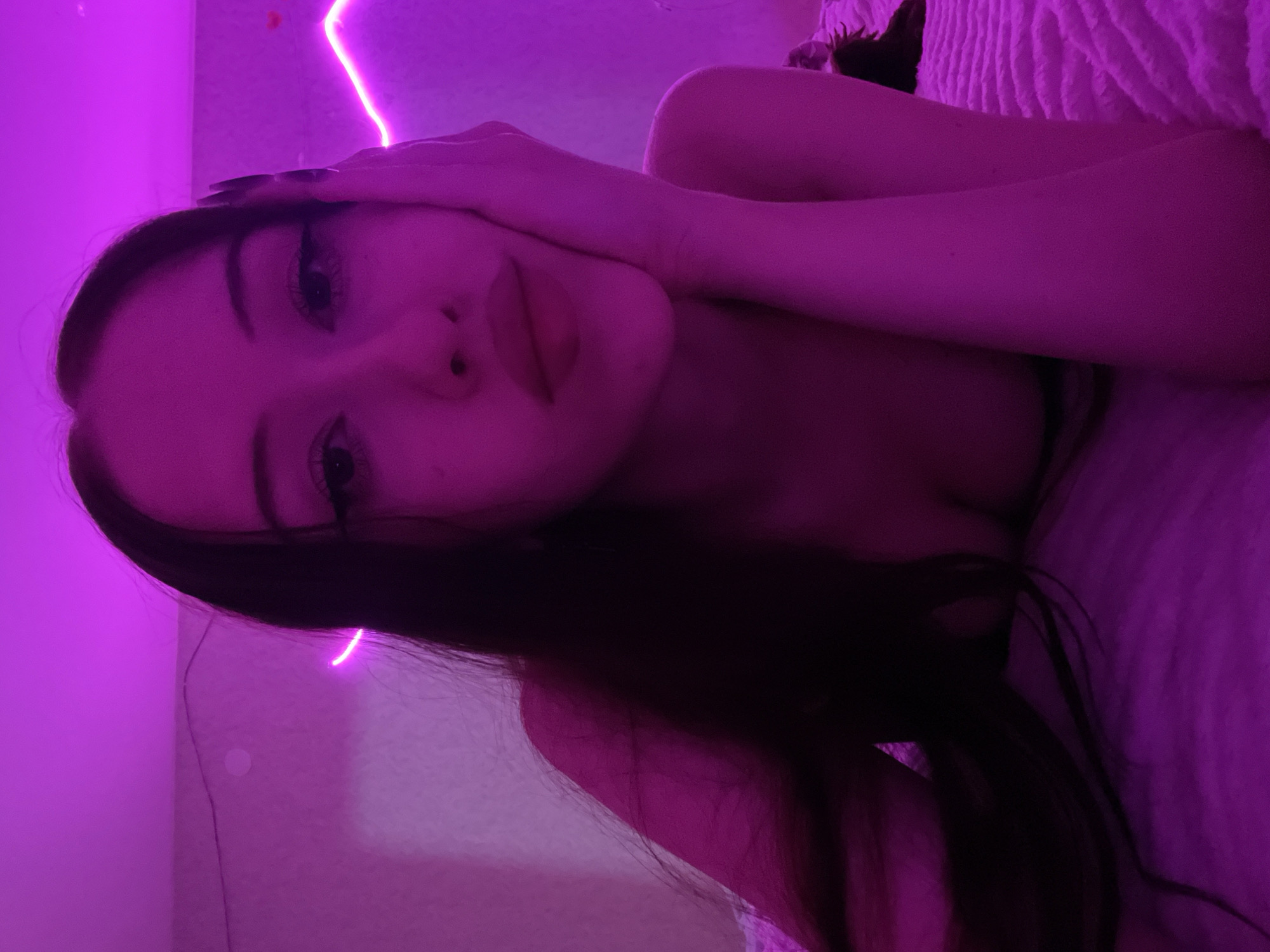 A young woman with long, dark hair lies on a bed bathed in purple light from neon strips. Her face is turned towards the camera, with her left hand resting on her cheek. She is wearing dark nail polish and appears to have winged eyeliner. The background is a textured wall with a faint circular light. The image is a selfie taken indoors. There is no information available to determine the location in Votkinsk, Russia.