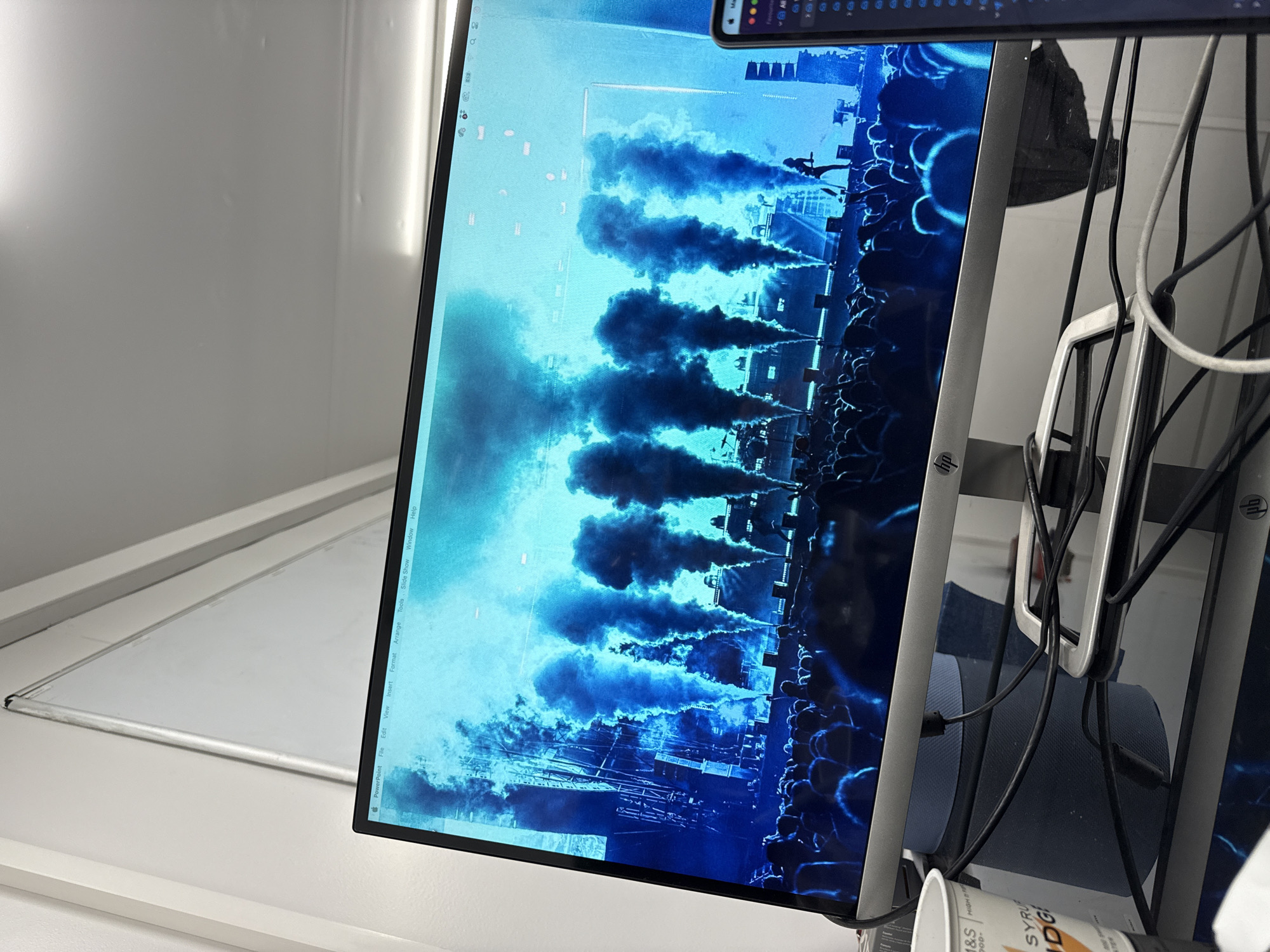 A monitor screen shows a blue-toned image appearing to depict a musical or theatrical performance, with multiple vertical plumes of dark smoke rising from a stage. A large crowd of indistinct figures is visible in the foreground. The monitor is situated in what appears to be an office or home setting, with part of a window frame visible to the left. To the right of the monitor, there are various cables and a partially visible white container with printed text. The image shown on the monitor appears to be a screenshot of a PowerPoint window, indicating that it might be a presentation or a slide currently being worked on. There are also the menu items File, Edit, View, Insert, Format, Arrange, Tools, Slide Show, Window, and Help showing on the top of the screen. The image does not provide sufficient information to determine a location in Epping Forest, United Kingdom.
