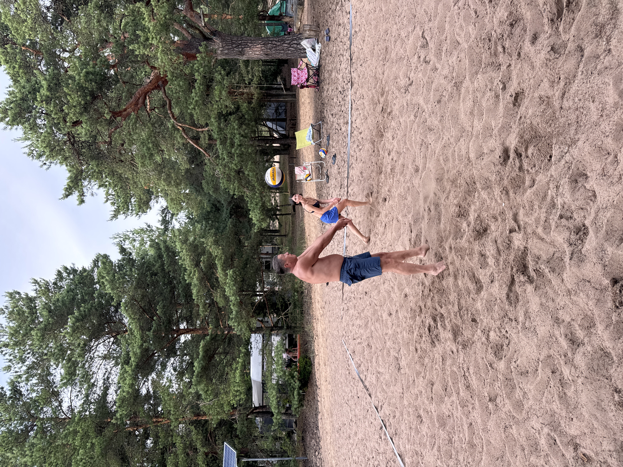 In Zelenaya Roshcha, Russia, two individuals are engaged in a game of beach volleyball on a sand court. A man in blue shorts stands in the foreground, his arms in position to receive or serve the ball. To his right, a woman in a black bikini top and blue shorts is in motion, her body angled towards the net, suggesting she is about to hit the volleyball that is suspended in the air above her. The court is marked with white lines, indicating the boundaries of the game. Surrounding the court is a natural environment characterized by tall pine trees with dense green foliage. In the background, RVs and tents are visible, hinting at a campsite. Several chairs, including a pink and a yellow one, are set up near the trees, along with a smaller volleyball and some footwear, suggesting a recreational area. The ground is covered in sand, with some footprints and uneven textures evident. The sky above is overcast, indicating a cloudy day.