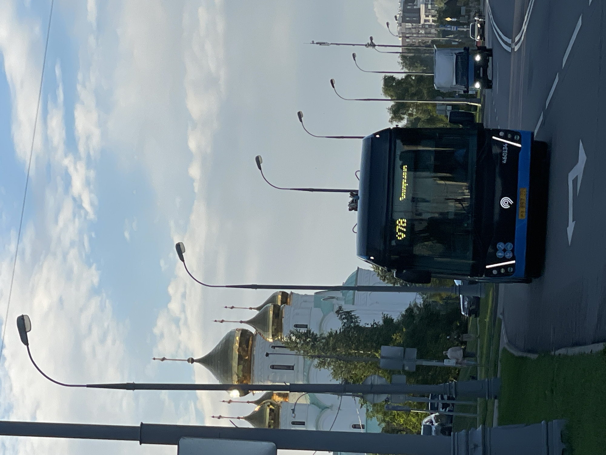 This is a low-angle, eye-level shot of a street scene in Moscow, Russia, featuring a modern blue and black trolleybus in the foreground. The trolleybus, displaying the route number "97B" and the text "Энтузиастов", is captured from the front as it moves along a multi-lane road. A silver truck with a covered bed is visible behind the trolleybus, followed by a yellow car. In the background, a large white building with multiple golden domes, characteristic of Russian Orthodox architecture, stands prominently against a partly cloudy sky. Several lampposts with curved arms are positioned along the roadside, illuminating the street. The scene depicts a typical urban environment with public transportation, vehicles, and a significant cultural landmark.