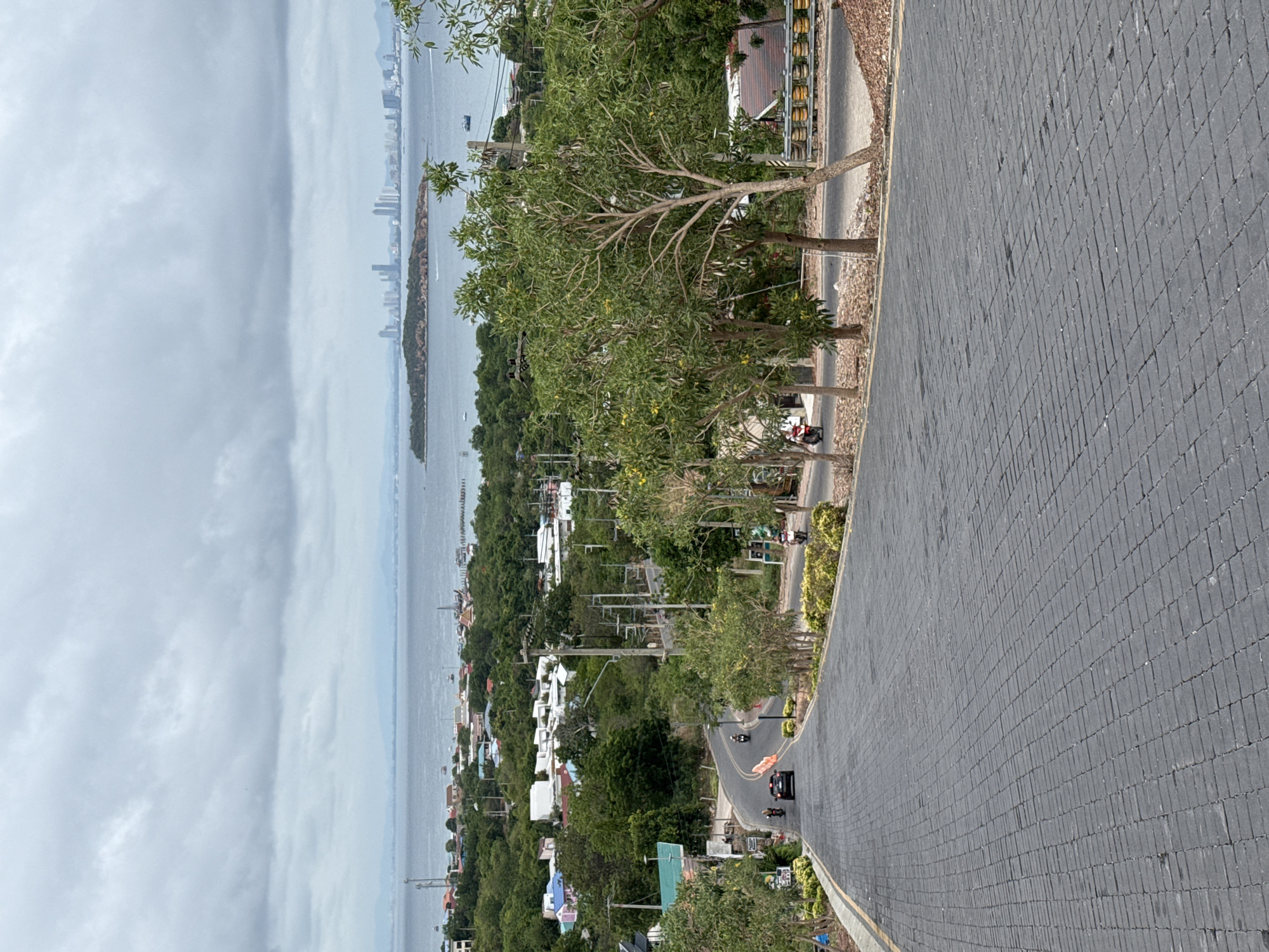 A high-angle view from a paved road in Pattaya City, Thailand, shows the sea, an island, and the city skyline under a cloudy sky. The road curves, with trees and buildings lining its side. Vehicles, including motorcycles and a car, are seen on the road.