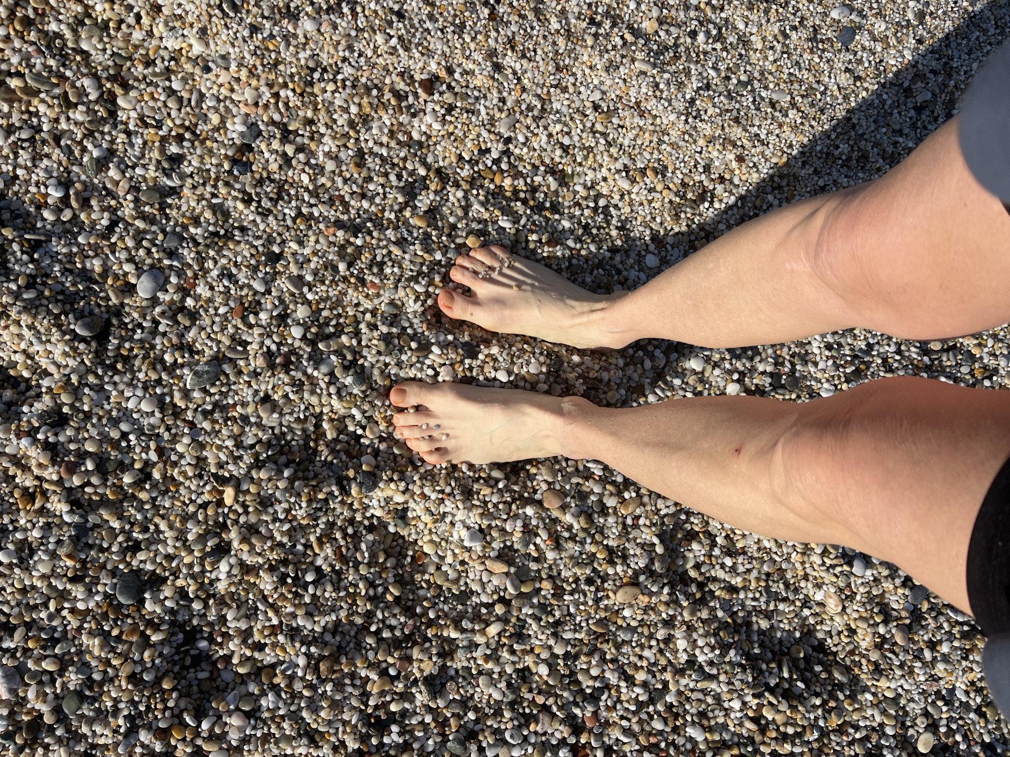 A high-angle shot shows a person's legs and feet standing on a pebble beach. The beach is composed of small, rounded pebbles of various colors, predominantly light brown, gray, and white. The person has fair skin and is wearing dark-colored shorts. The legs and feet are positioned in the center of the frame, with the feet angled slightly outwards. The background is entirely composed of the pebble beach, which extends beyond the visible frame. The lighting appears to be natural daylight, casting shadows from the legs and feet onto the pebbles. Based on the landscape, the image can be placed at a beach in Alanya, Turkey.