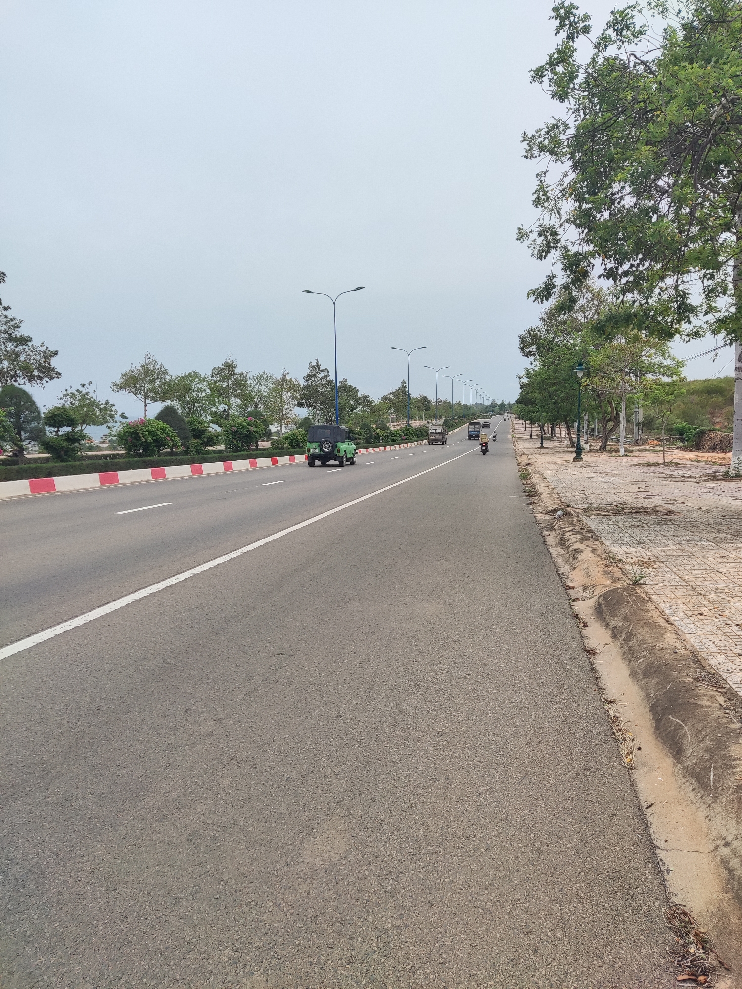 The image captures a wide, paved road stretching into the distance, with a white line dividing the lanes. A green, open-top vehicle is visible in the lane closest to the viewer, moving in the same direction as the perspective. Further down the road, other vehicles including what looks like a truck and a smaller motorcycle are also visible.
To the left of the road is a red and white striped barrier, behind which lies a line of trees and bushes. To the right of the road, a paved sidewalk runs parallel, lined with trees and streetlights. The pavement is cracked and somewhat uneven. Beyond the sidewalk, the land slopes upward, covered in sparse vegetation.
The sky above is overcast, suggesting a cloudy day. The overall scene is a street view within a city in Unknown, Unknown, depicting typical road infrastructure alongside natural elements.