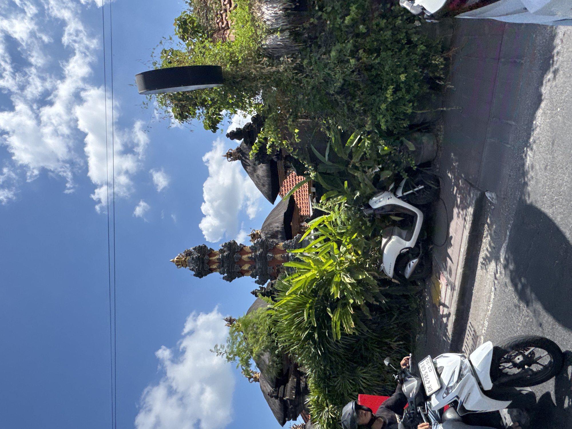 The image captures a vibrant street scene in North Kuta, Indonesia, on a bright, sunny day with a blue sky and scattered white clouds. The setting features traditional Balinese architecture and lush tropical vegetation alongside a paved road.
In the foreground, a person wearing a black helmet and dark clothing is visible on a white Honda scooter, partially obscuring its front. The scooter's license plate, "DK 4785 GBT," with an expiry of "03-26," is clearly visible. Another white scooter, possibly a Honda Scoopy, is parked further down the street on the right. The paved road features a narrow concrete curb separating it from the greenery, and shadows are cast across the asphalt, indicating strong overhead sunlight.
To the left, a prominent traditional Balinese temple or shrine with an elaborately decorated, towering spire, adorned with gold and grey elements, stands out against the sky. Adjacent to it, traditional buildings with thatched roofs are nestled among dense, green foliage, including palm-like plants and various bushes. A black, circular object, possibly a street light or sign, is partially visible above the greenery on the far left. Power lines stretch across the blue sky. The scene depicts a typical day with local transport and distinctive cultural landmarks, reflecting the characteristic environment of Bali.
