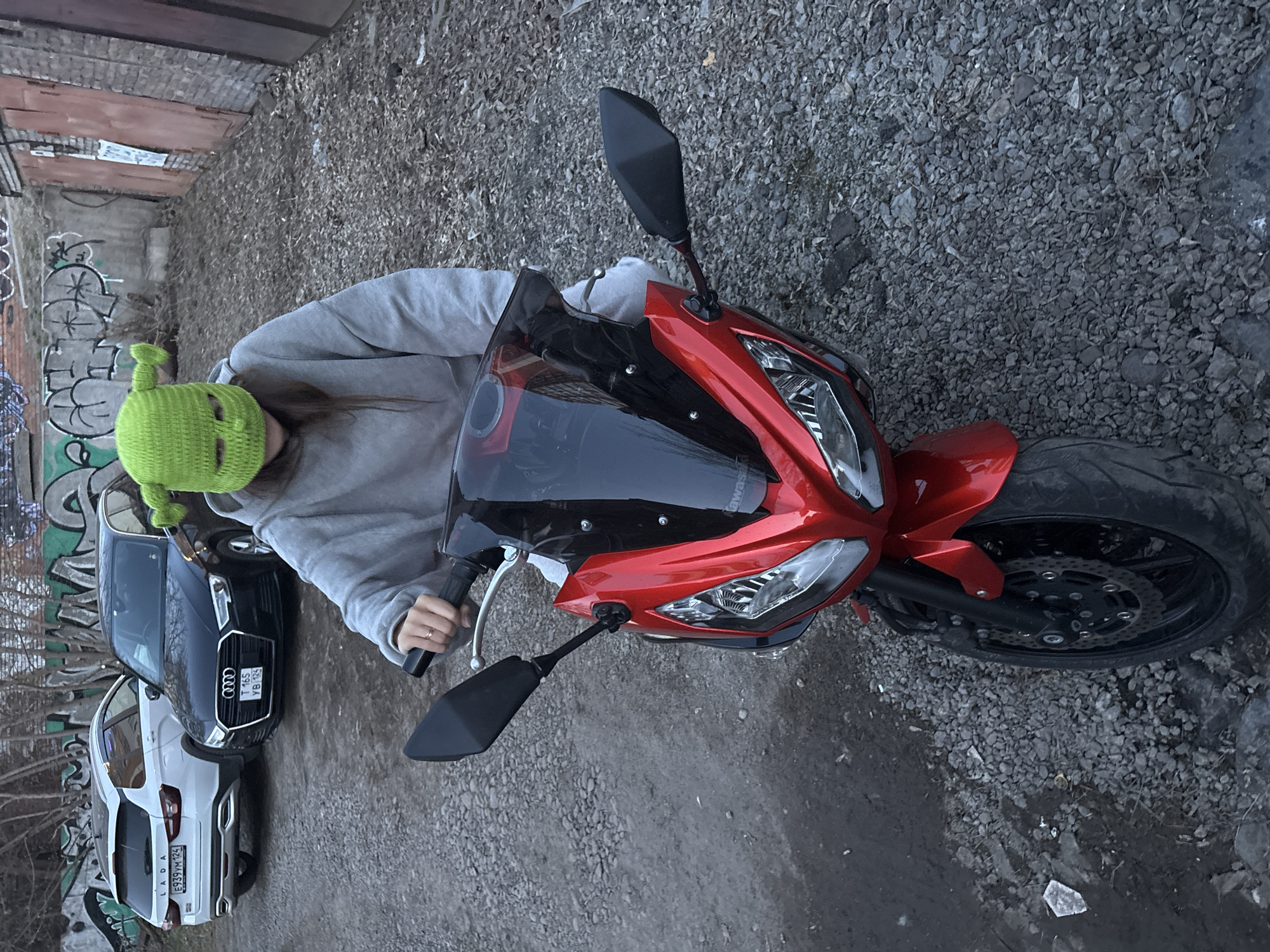The photograph depicts a person wearing a green, crocheted balaclava on a red motorcycle, with their hands on the handlebars. The motorcycle is parked on a gravel surface. Behind the motorcycle, there are two parked cars: a white Lada and a black Audi. The license plate of the Lada reads "E939WM124". The Audi's license plate reads "T16SYB124". In the background, there's a wall covered with graffiti. Given the license plate code "124," which is assigned to the Krasnoyarsk region of Russia, the location is likely within the city of Krasnoyarsk, Russia. The environment seems to be an alleyway or a parking area with signs of disrepair and graffiti, suggesting an urban setting.