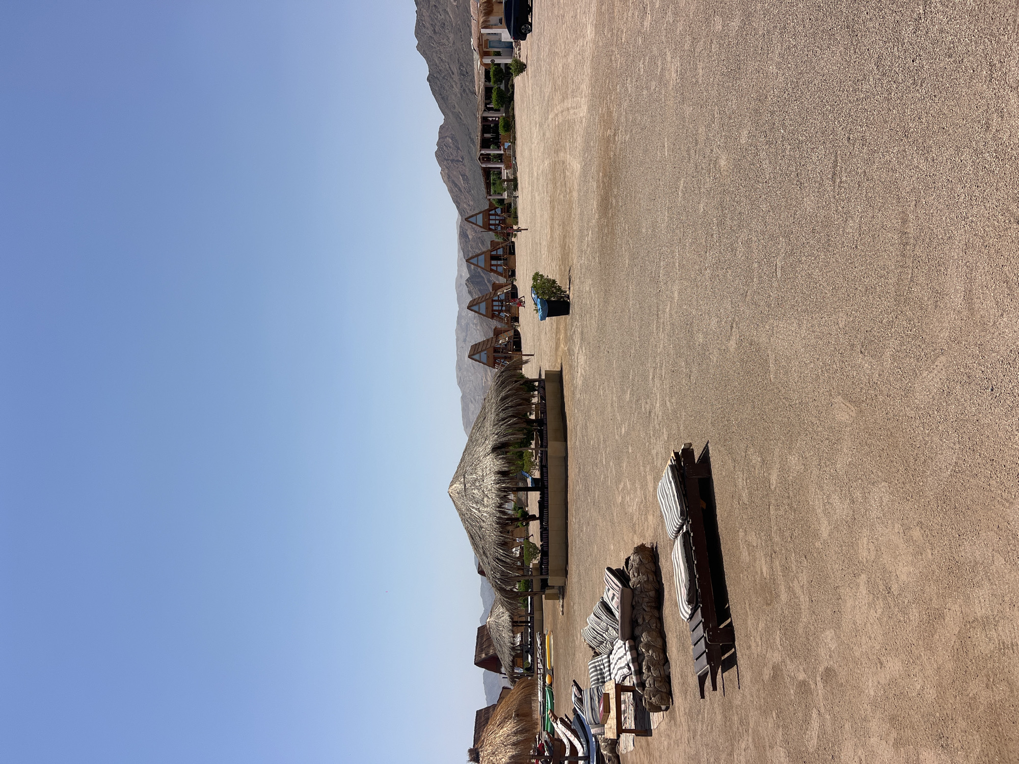 The image shows a resort area in Nueiba, Egypt, on a clear, sunny day. In the foreground, there are several lounge chairs with striped cushions on a sandy or gravelly ground. To the left, there is a low seating area with cushions and a small table. Behind this, there are more thatched-roof structures, including a large, round hut providing shade. In the middle ground, a row of distinctive triangular cabins with wooden exteriors and dark roofs are visible, set against a backdrop of arid, mountainous terrain. Further back, a low-rise building with a light-colored facade and a dark roof appears to be part of the resort. A black car is parked on the right side of the frame. A blue trash bin is situated near the triangular cabins. The sky is a uniform, light blue. The scene conveys a sense of a quiet, outdoor leisure destination in a desert environment near the coast.