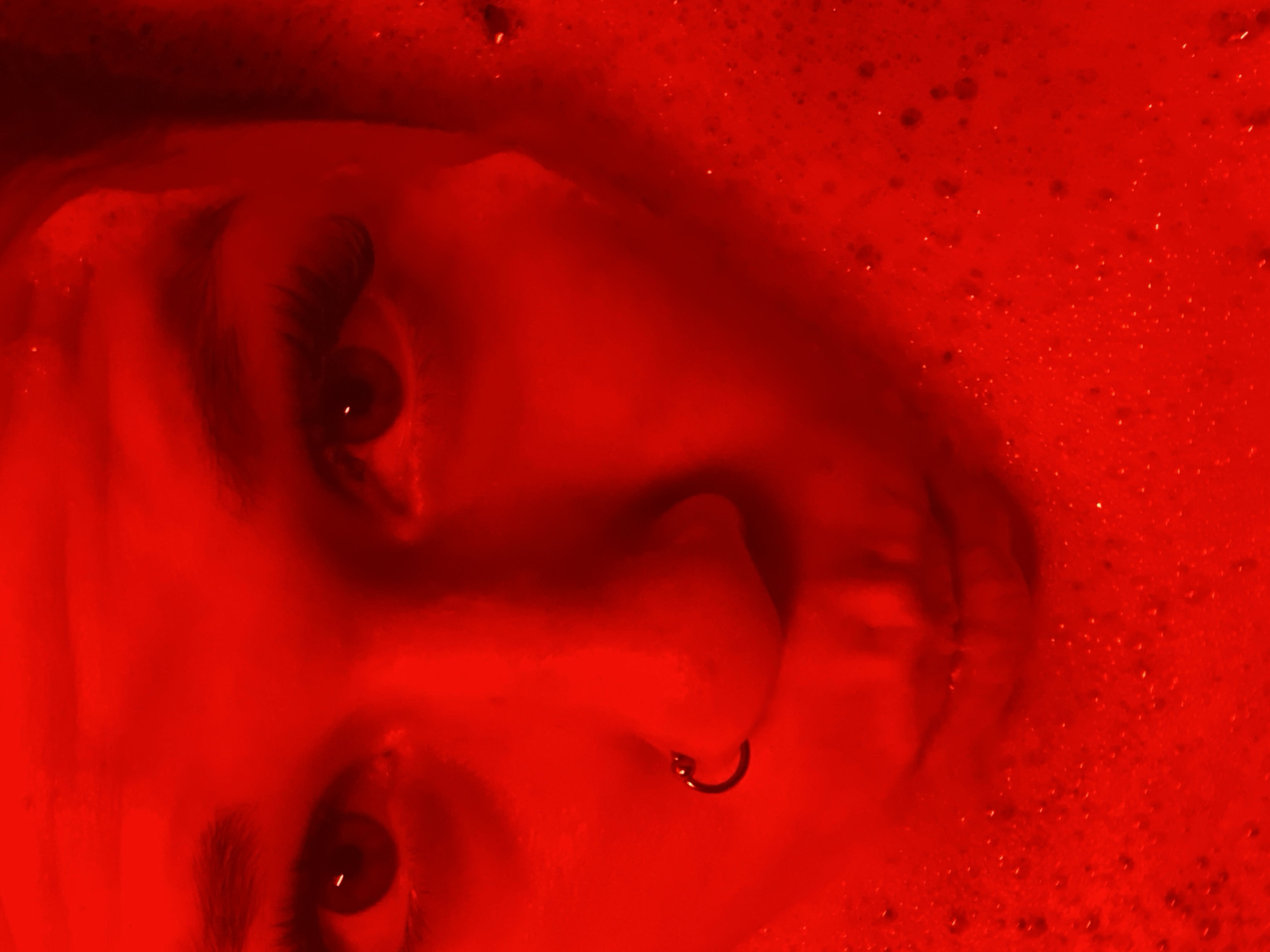 The image shows a close-up of a person's face, illuminated with a red light. The person has dark eyebrows and eyelashes, and a nose ring. The background is filled with red bubbles, suggesting the person may be in a bath or pool. The location in the city of Chaikovsky, Russia is not discernible from the image.