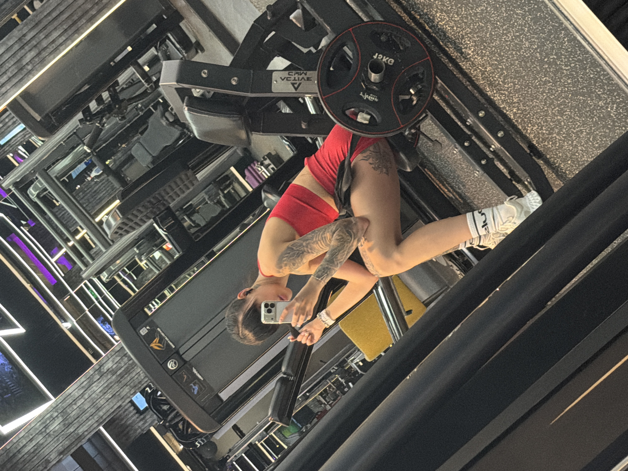A woman with tattoos on her arms and legs is taking a selfie in a gym. She is wearing a red sports bra and red shorts, and white socks with white athletic shoes. She is sitting on a weight machine with a weight plate visible. The gym appears to be modern with exposed ceilings and neon lighting. The image does not provide enough information to determine the specific location within the city of Plovdiv, Bulgaria.