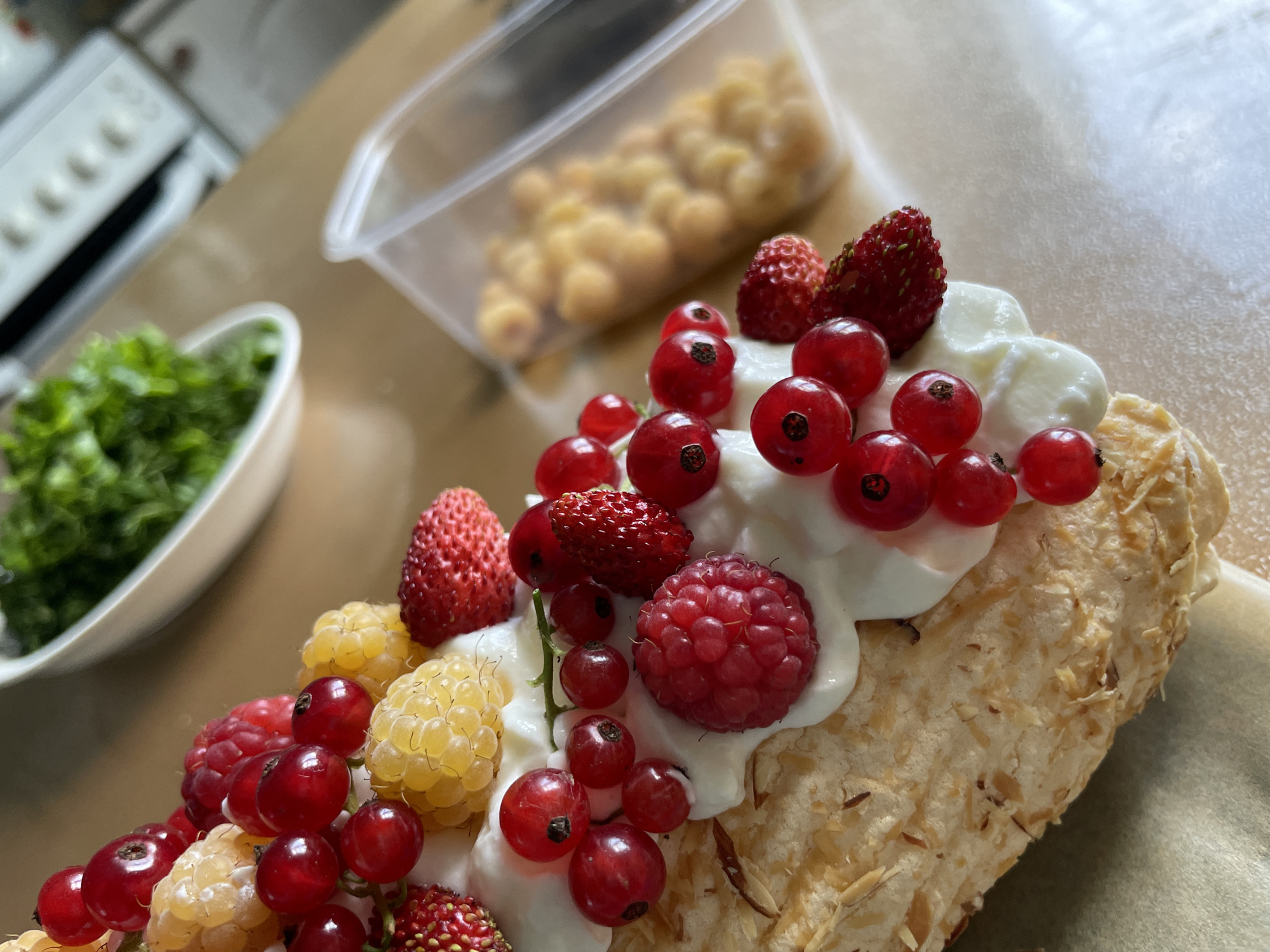 In a close-up shot, a dessert is displayed on a light-colored surface. The dessert appears to be a meringue base, sprinkled with slivered almonds, topped with a dollop of white cream. Adorning the cream are an assortment of fresh berries, including bright red currants, plump red raspberries, and small, yellow raspberries. There are also a few tiny wild strawberries scattered amongst the larger berries. In the background, slightly out of focus, is a white bowl filled with chopped green herbs, and a clear plastic container holding more yellow berries. The scene suggests a preparation or serving of a sweet treat, likely in a domestic setting, as indicated by the glimpse of kitchen appliances in the upper right background. The location is described as Дресвяново, Russia, though there are no specific landmarks in the image to confirm this or any indications of urban life beyond what might be found in a home.