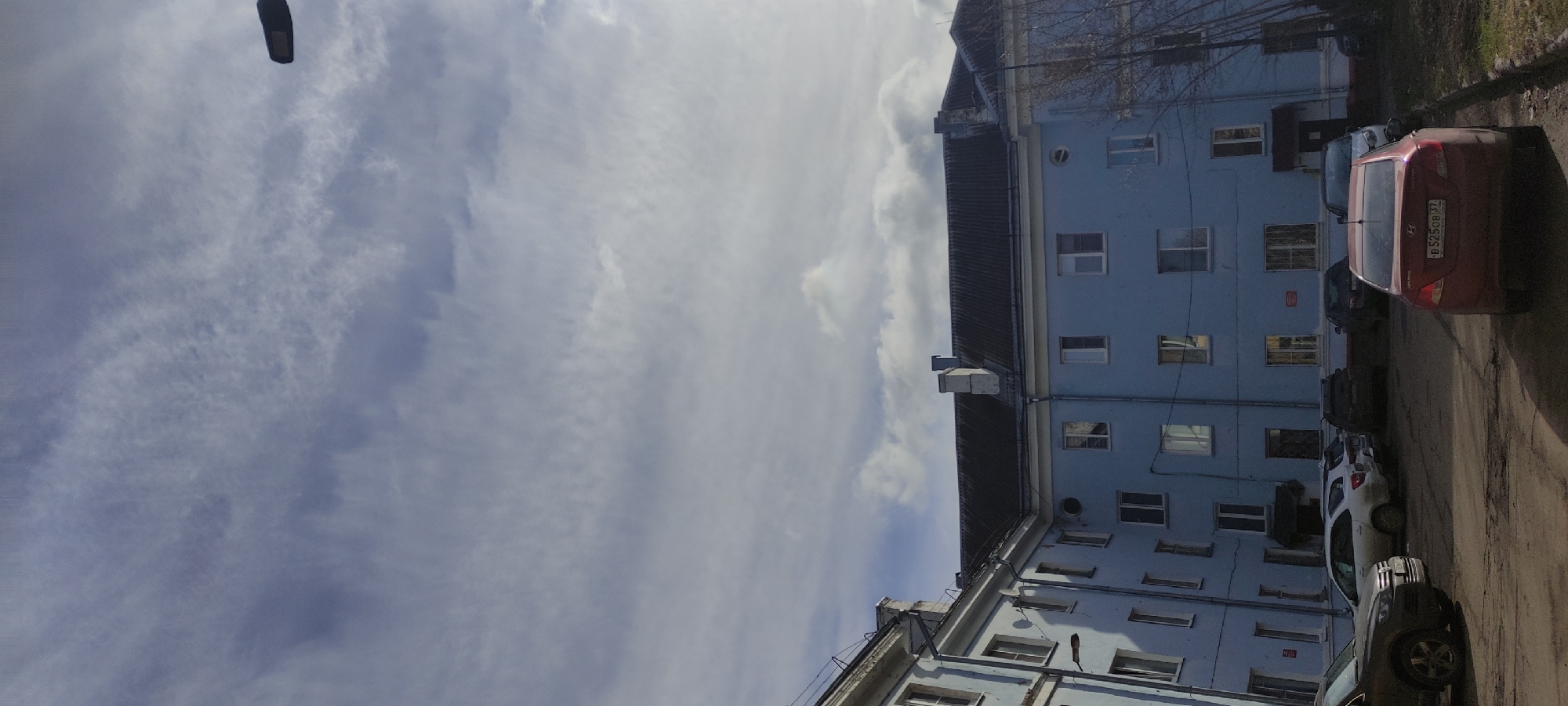 The image depicts a blue-painted multi-story building on the right side, with several parked cars along the side of the building. The sky occupies most of the left side of the image, displaying light and wispy clouds. A street lamp is visible at the top left corner. The location is Fryazino, Russia, as indicated by the car license plate number "B 525 OB 32" on a red car which corresponds to Bryansk Oblast, Russia. The overall environment suggests a residential or urban area.