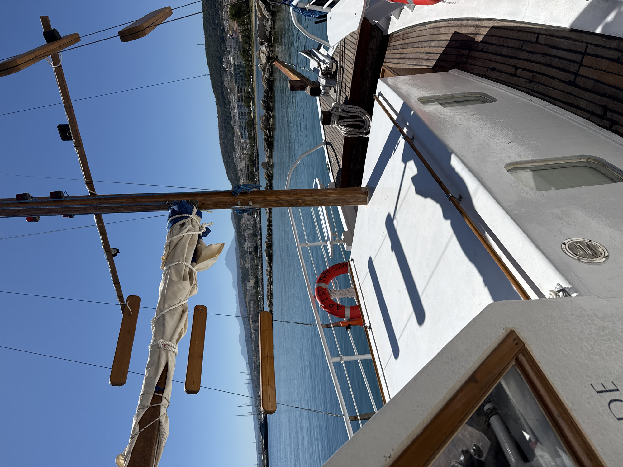 A sunlit, low-angle, full outdoor shot captures a view from the deck of a white boat, likely a traditional wooden sailing vessel, anchored in the clear, blue waters of Rethymno, Greece. The boat's white deck dominates the foreground, with a wooden trim and a portion of a glass-covered helm visible on the left. To the right, a wooden deck runs along the boat's side, transitioning into a white superstructure with several rectangular windows. A red life preserver with Greek lettering is affixed to the white railing.
The middle ground shows the boat's mast, a rolled-up sail, and rigging, along with a wooden boom extending horizontally. Wooden steps or planks hang suspended from the rigging. Beyond the boat, the calm, blue sea stretches towards a distant coastline. The coastline is lined with buildings, ranging from white to pastel-colored structures, nestled against a green, hilly landscape. Several sailboats with tall masts are visible further in the harbor, indicating a populated marina. The sky is a clear, vibrant blue, with distant mountains forming a hazy backdrop. The overall scene is tranquil and suggests a typical sunny day in a coastal Greek town.