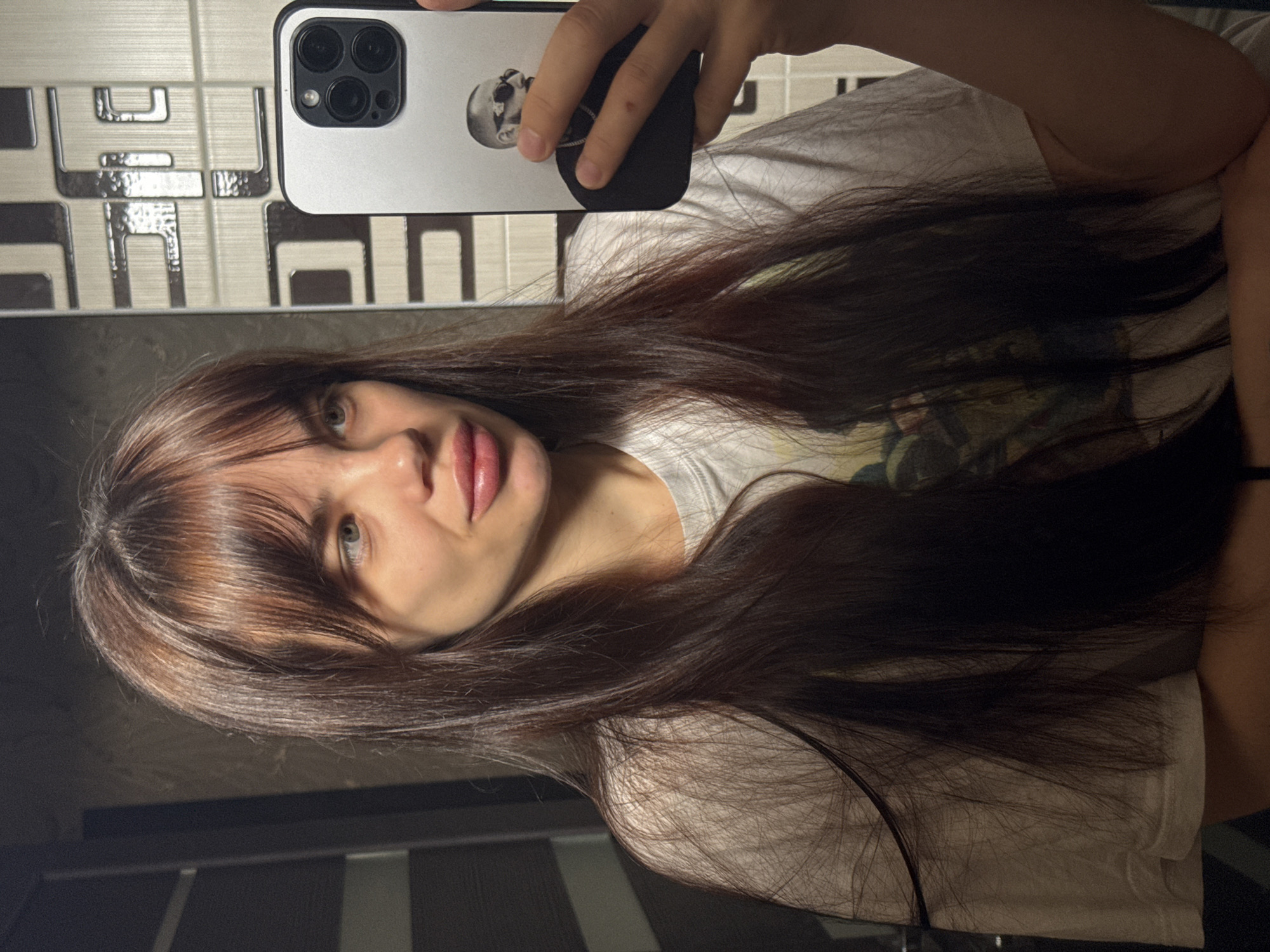 A young woman with long, dark hair and bangs is taking a selfie with a smartphone. The phone has a white case with a graphic of a skull. She is wearing a white t-shirt. The background is a patterned tile wall. It is not possible to ascertain if this is Rostov-on-Don, Russia.