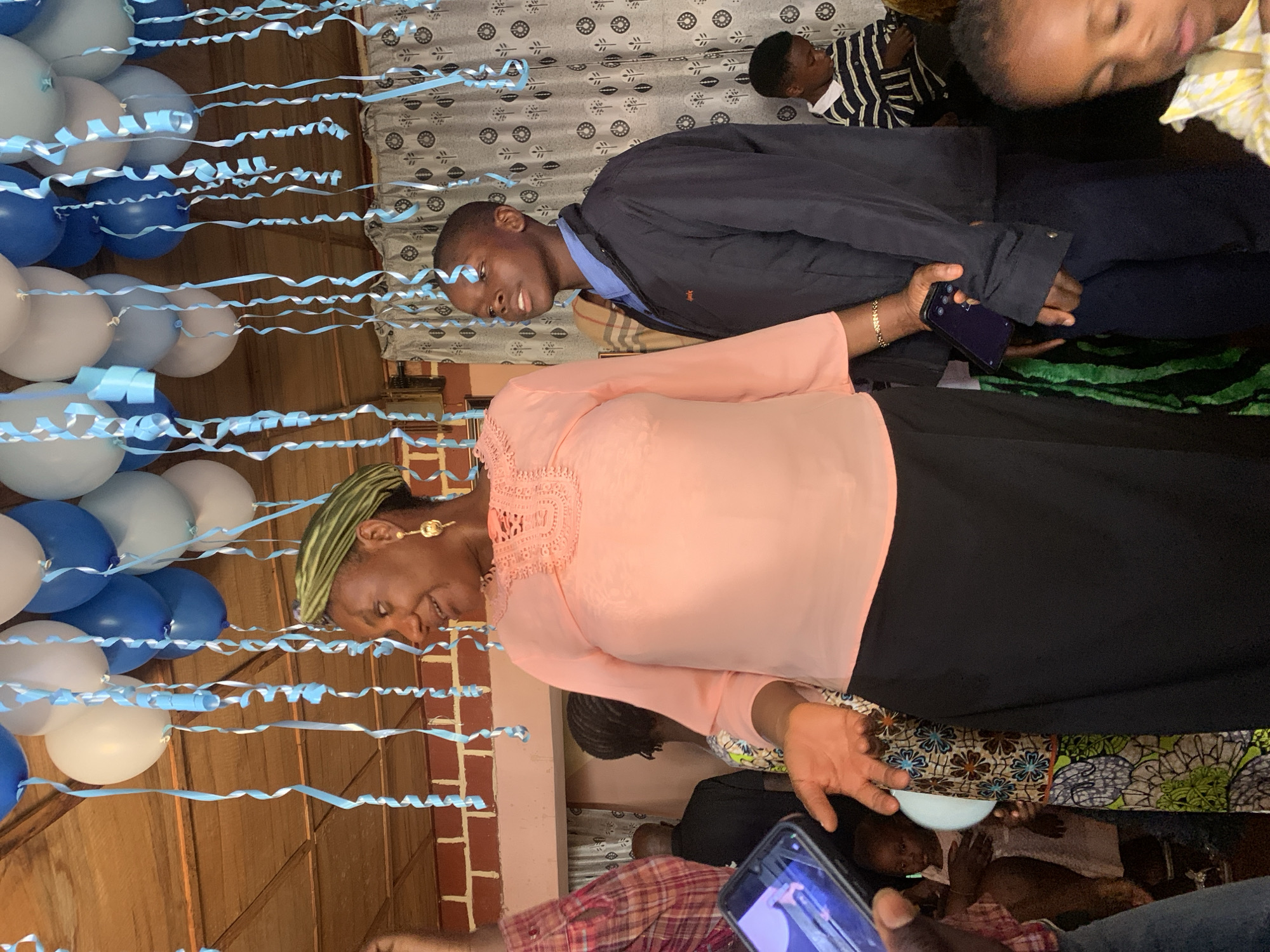 A photograph depicts an indoor celebratory event, potentially in the city of Jos, Nigeria, though no landmarks are visible in the image to confirm the location. Balloons and streamers adorn the space. An adult woman with a light peach top and black skirt, wearing a green head covering, is standing. A younger man in a blue shirt and a dark jacket stands next to her. An individual in a striped shirt and another person in a yellow top appear in the background. There are brick structures behind the balloons and a white ceramic bowl. Someone is recording a video with a mobile phone.