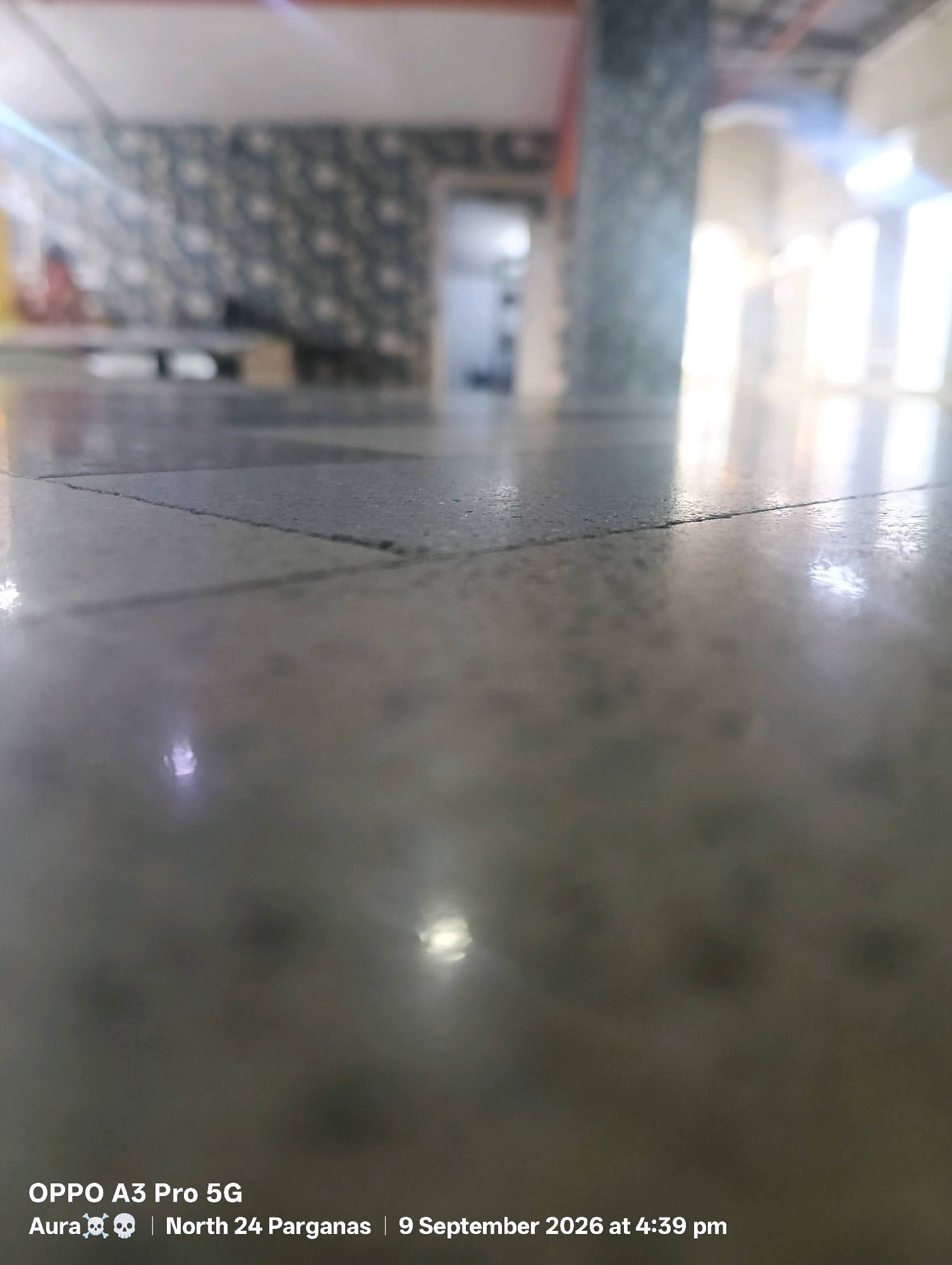 The image shows a low-angle shot of a polished concrete floor in what appears to be an indoor public space. The floor is tiled with large, square tiles, and there is a prominent crack running across one of the tiles in the foreground. Light reflects off the smooth surface of the floor, creating bright highlights. In the background, the scene is blurred, but it is possible to discern architectural elements such as pillars and windows with bright light streaming through. There is also a glimpse of patterned wallpaper on a wall. Text overlaid on the bottom of the image indicates the location as "North 24 Parganas" and the date as "9 September 2026 at 4:39 pm". Given that North 24 Parganas is a district in West Bengal, India, and Barasat is the administrative headquarters of this district, it is plausible the image was taken in Barasat. The image also contains watermarks indicating "OPPO A3 Pro 5G" and "Aura 💀💀". No people are clearly visible or discernible in the image.