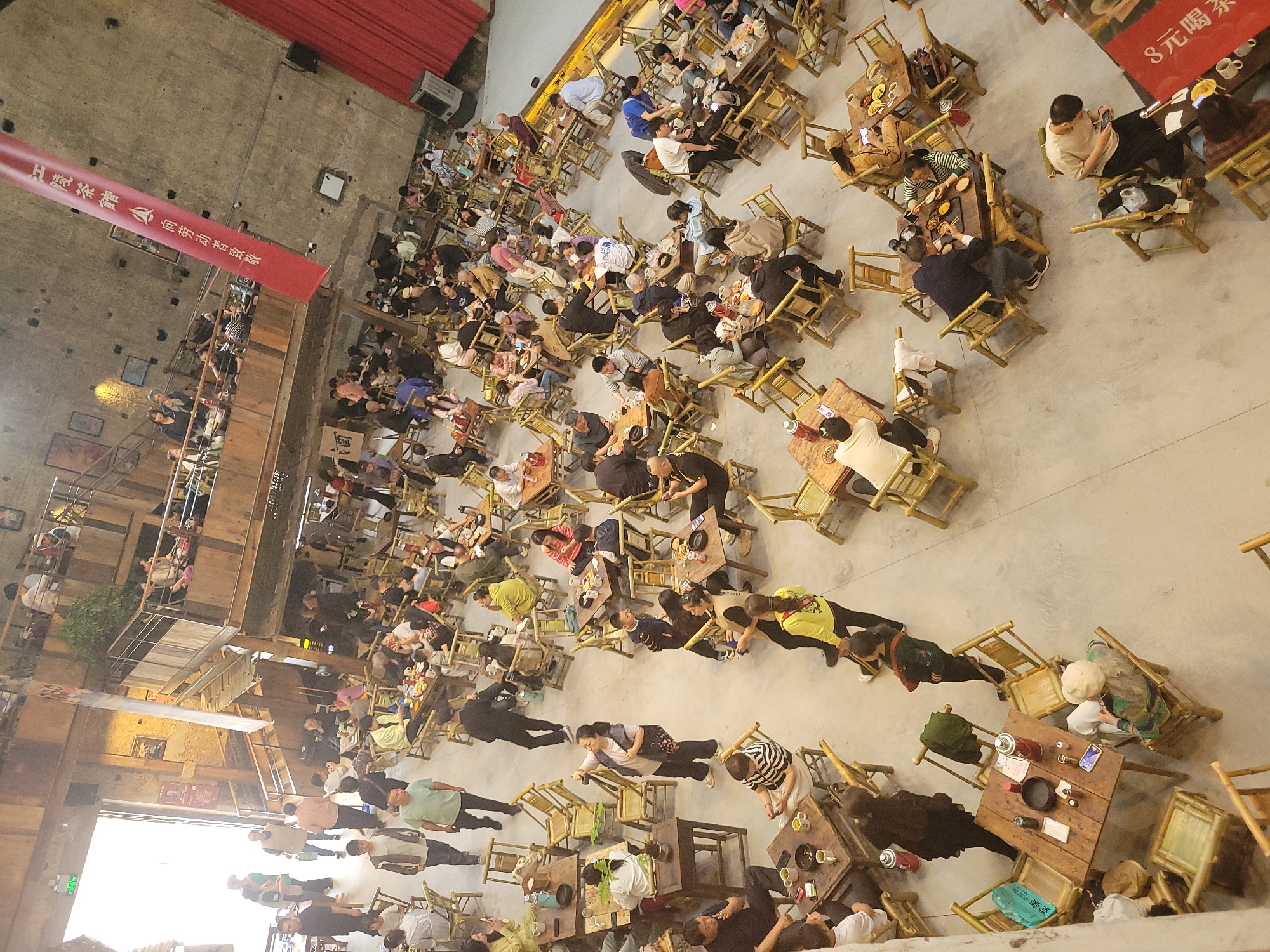 A high-angle shot shows a crowded outdoor venue in Liangjiang, China, featuring a large gathering of people seated at numerous small wooden tables and chairs. Most tables are occupied by groups of people eating and drinking, and some people are walking between the tables. The ground is a light-colored, smooth surface. The venue is enclosed by tall buildings and a red curtain on one side. A red banner with white Chinese characters hangs from the ceiling. A multi-level building with wooden railings and stairs is visible on the left side of the image.