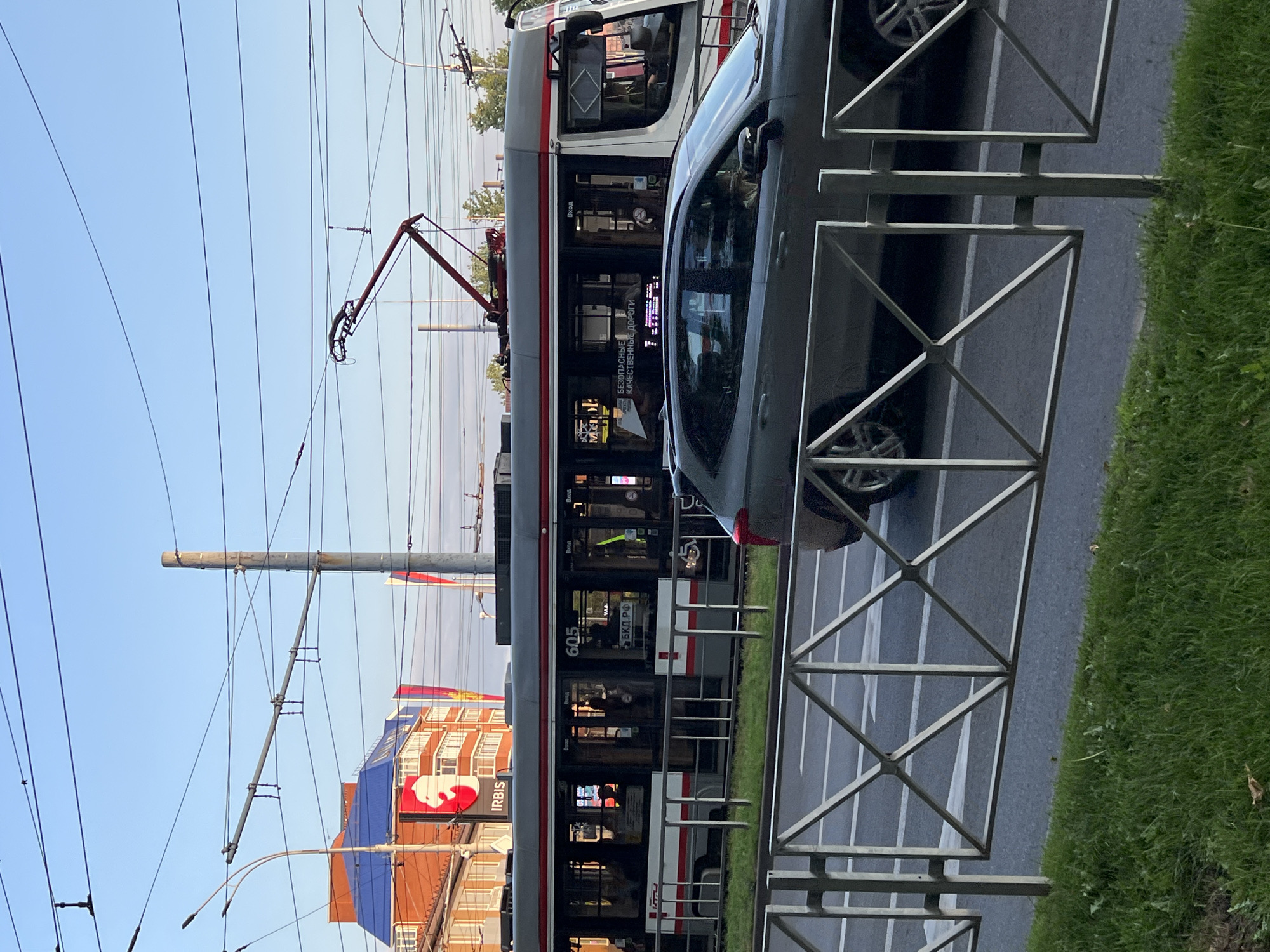 The image shows a street scene in Krasnodar, Russia, with a tram and a car. The tram, identified by the number 605, is gray with red and white trim and is moving along the tracks. Inside the tram, several passengers are visible through the windows. A gray SUV is driving on the road next to the tram. A metal fence separates the sidewalk and grassy area from the street. In the background, there is a building with orange brick and a blue roof, and a large red sign with a white symbol and the word "IRBIS." Overhead, there are numerous electrical wires for the tram. The sky is clear and blue, indicating it is daytime.