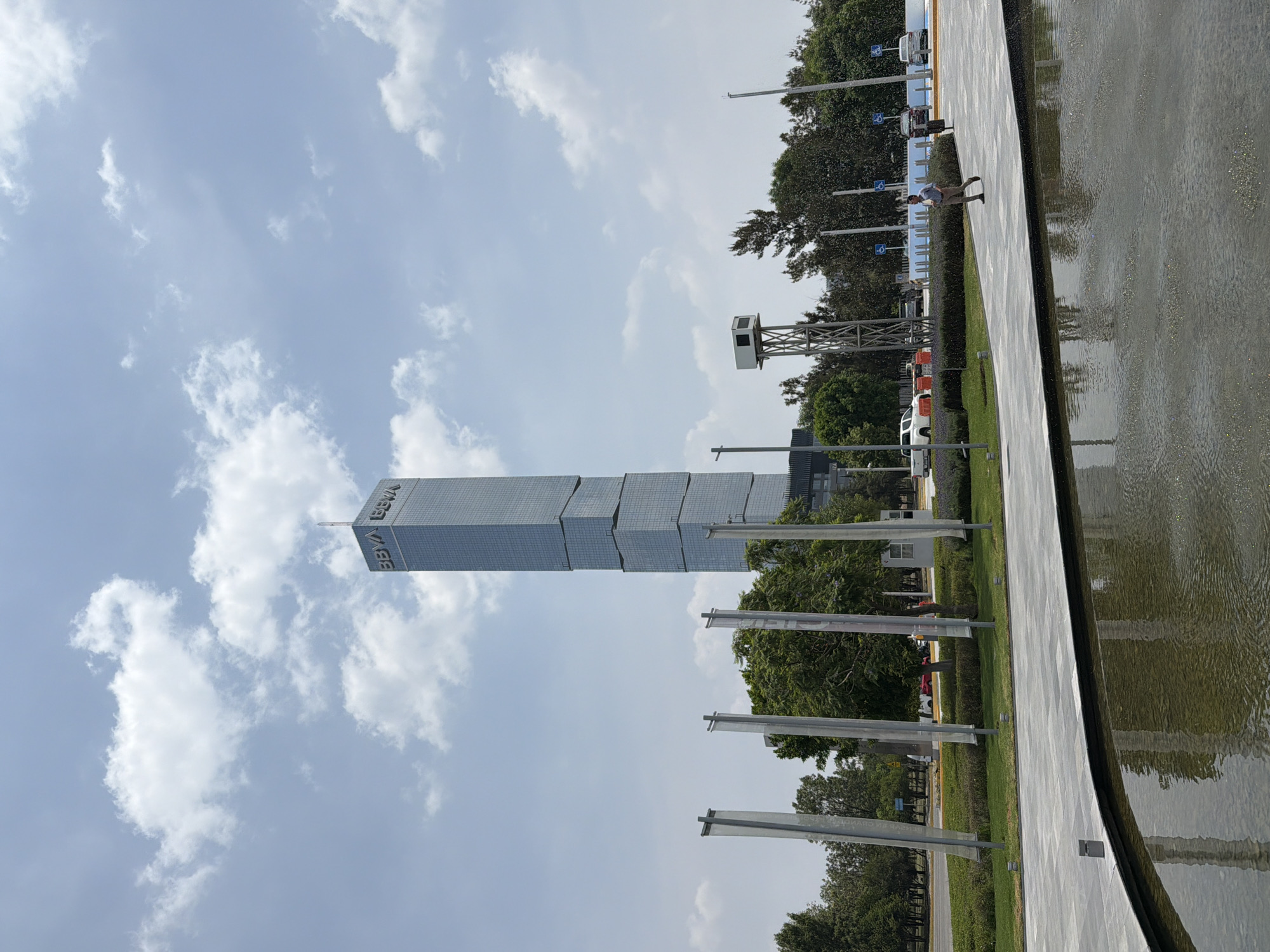 The image shows the BBVA Tower in San Bernardino Tlaxcalancingo, Mexico, under a sky with scattered clouds. The tower appears to be constructed from stacked, rectangular blocks. In the foreground, there's a reflective pool and a concrete walkway. Along the walkway are flagpoles and greenery. People can be seen walking along the path near the pool. In the background, trees and a small observation tower are visible. Various vehicles can be seen parked along a road behind the trees.
