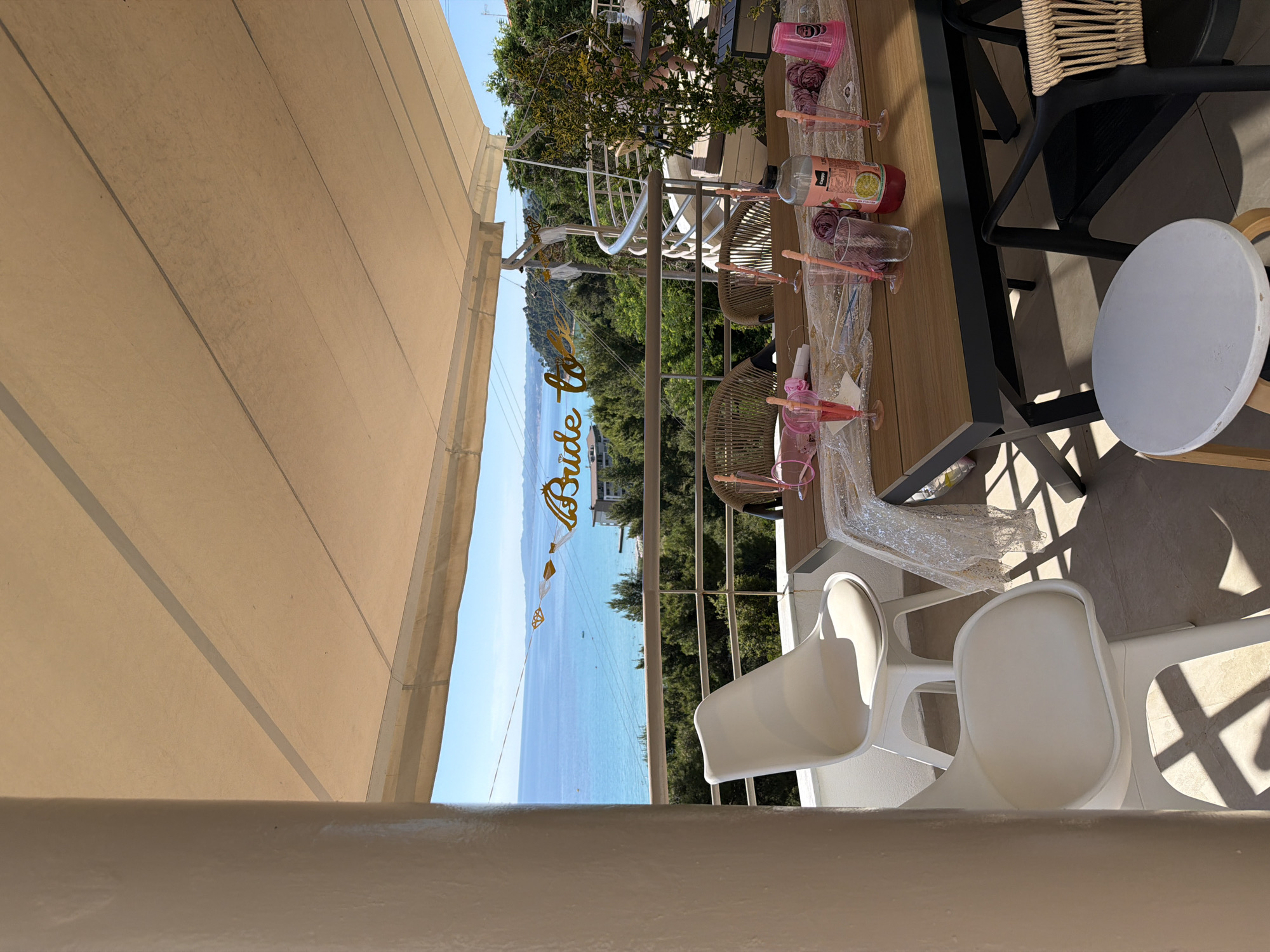 The image shows a sun-drenched balcony in the city of Kassandra Municipal Unit, Greece, offering a breathtaking view of the Aegean Sea. A beige awning extends over the balcony, providing shade. A white pillar stands to the left, casting a shadow on the tiled floor. In the foreground, a wooden table is set for a celebration, adorned with a sequined runner, pink plastic cups, glassware, and decorative roses. A bottle of pink beverage sits on the table. Two white chairs, one with a cushion, are positioned at the table. In the background, a banner that reads "Bride to be" is hung across the railing, with decorative tassels. Beyond the balcony, lush green trees and buildings dot the landscape leading down to the turquoise sea, which stretches towards a distant horizon under a clear blue sky. Several chairs, including two wicker chairs, are visible on the balcony.