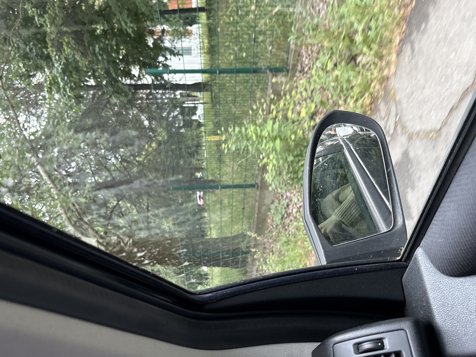 The media file captures a view from inside a vehicle, looking outwards through a side window and a partial view of what appears to be a sunroof or open roof. The setting is an outdoor, green environment, likely a roadside or park area within the Cukarica Urban Municipality, Serbia.
In the immediate foreground, parts of the car's interior are visible, including a dark door frame, window seals, and a control dial on the dashboard. A prominent black side mirror on the right reflects a portion of the vehicle's exterior and a ribbed fabric, possibly from a seat or clothing.
Beyond the vehicle, a green wire or chain-link fence runs horizontally across the scene, separating lush green vegetation in the foreground from a dense canopy of trees and bushes in the background. A paved path or road is visible on the right side, extending into the distance alongside the fence. A small, indistinct red and white object can be seen hanging or placed near the fence in the mid-ground. Faintly in the distance behind the fence, a structure, possibly a building, is visible.
The lighting suggests it is daytime, with diffused light indicating an overcast sky or soft sunlight. There are no strong shadows, and the weather appears calm. No people are present in the image, and no visible text can be discerned.